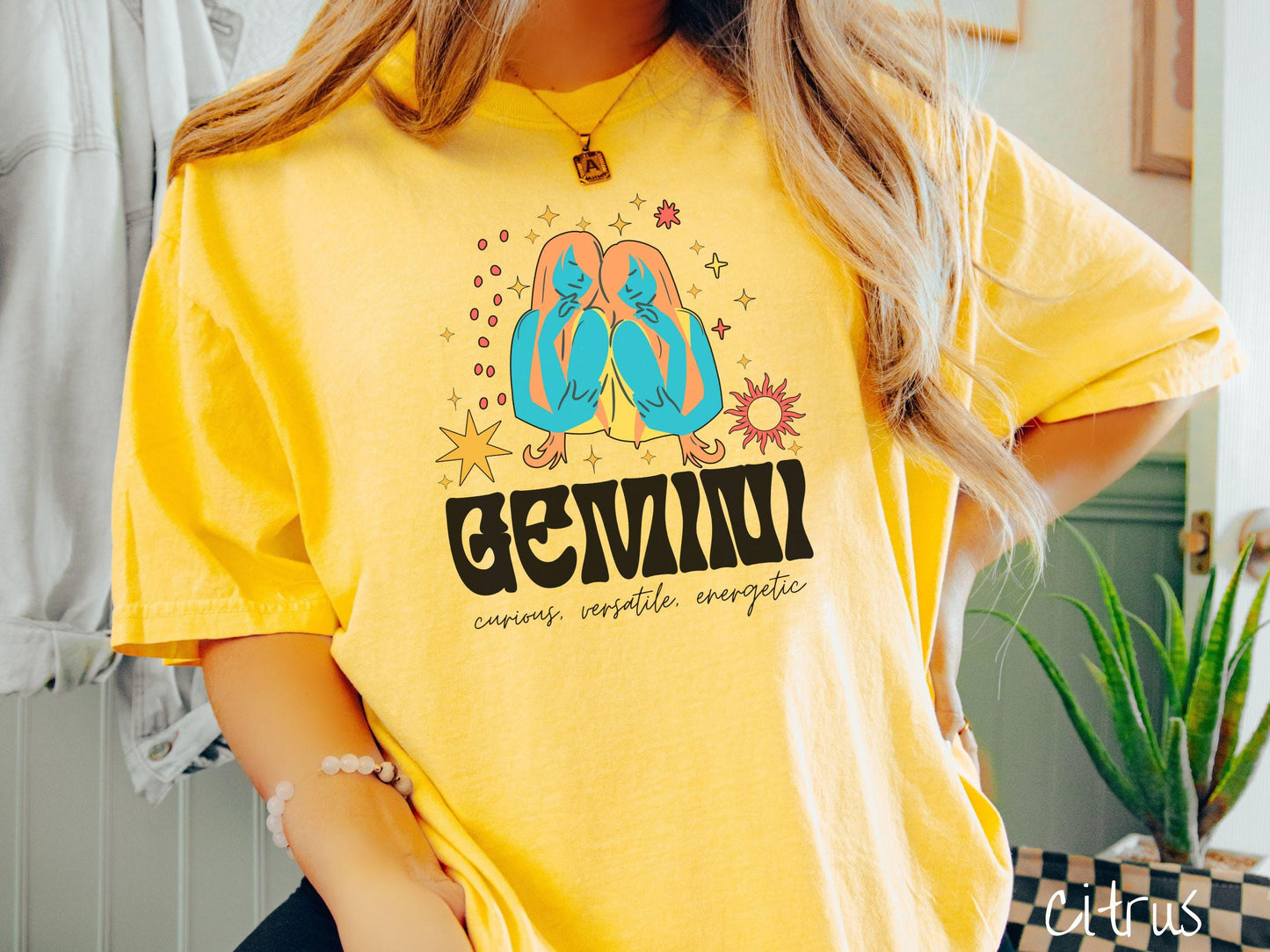 A vintage citrus colored Comfort Colors T-shirt features the twins Castor and Pollux in blue and orange colors surrounded by stars and the sun with text Gemini in large bold font and below are the words curious, versatile, energetic in cursive font.