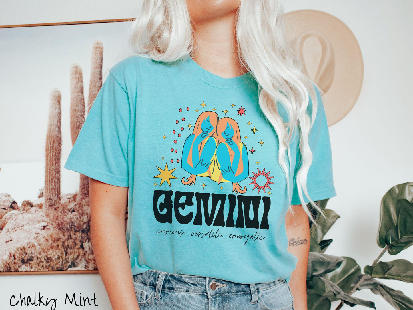 A vintage chalky mint colored Comfort Colors T-shirt features the twins Castor and Pollux in blue and orange colors by stars and the sun with text Gemini in large bold font and below are the words curious, versatile, energetic in cursive font.