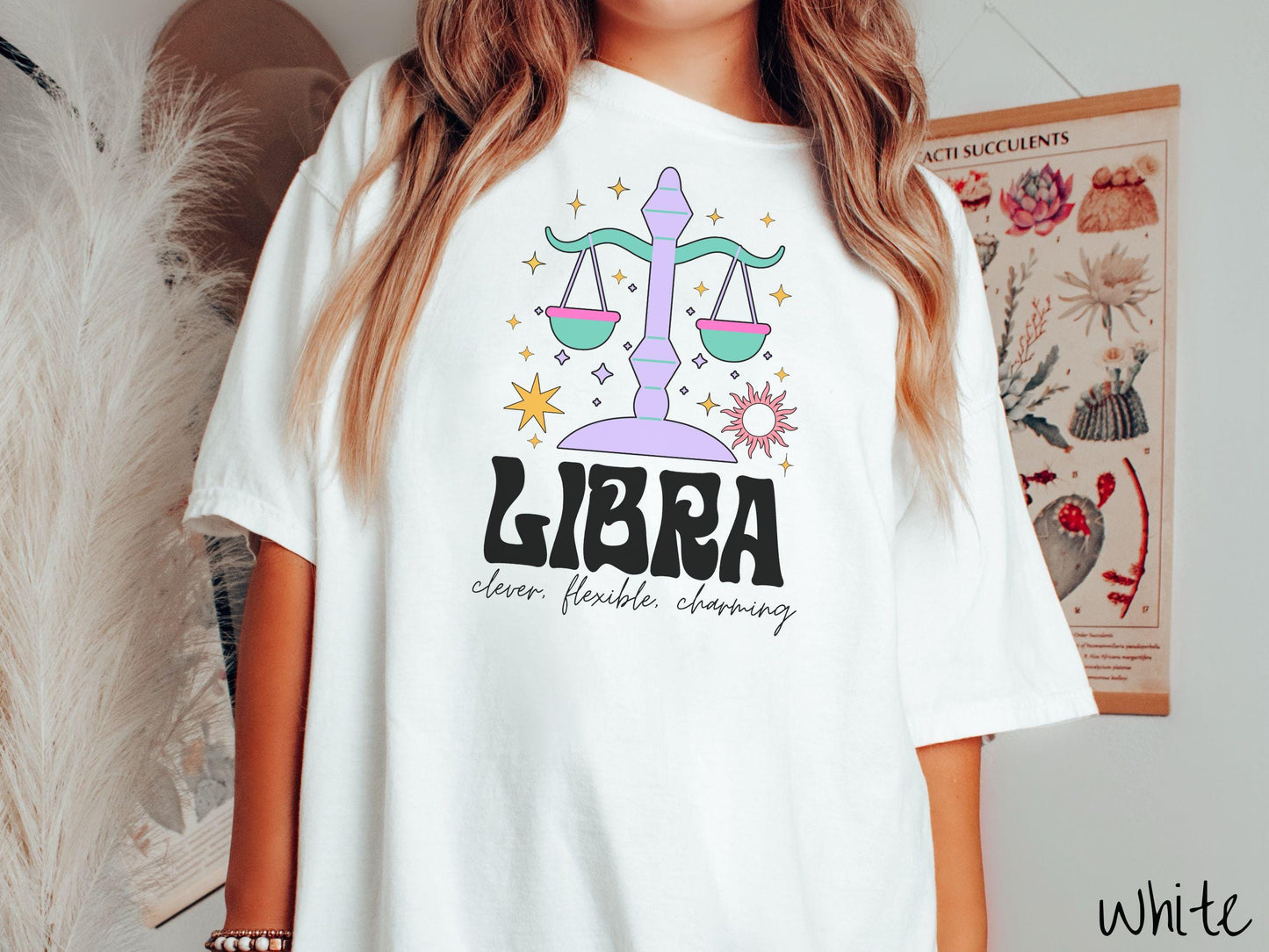 A vintage white colored Comfort Colors T-shirt features a large purple and green weight scale surrounded by stars and the sun with the text Libra in large bold font and below that are the words clever, flexible, charming in cursive font.