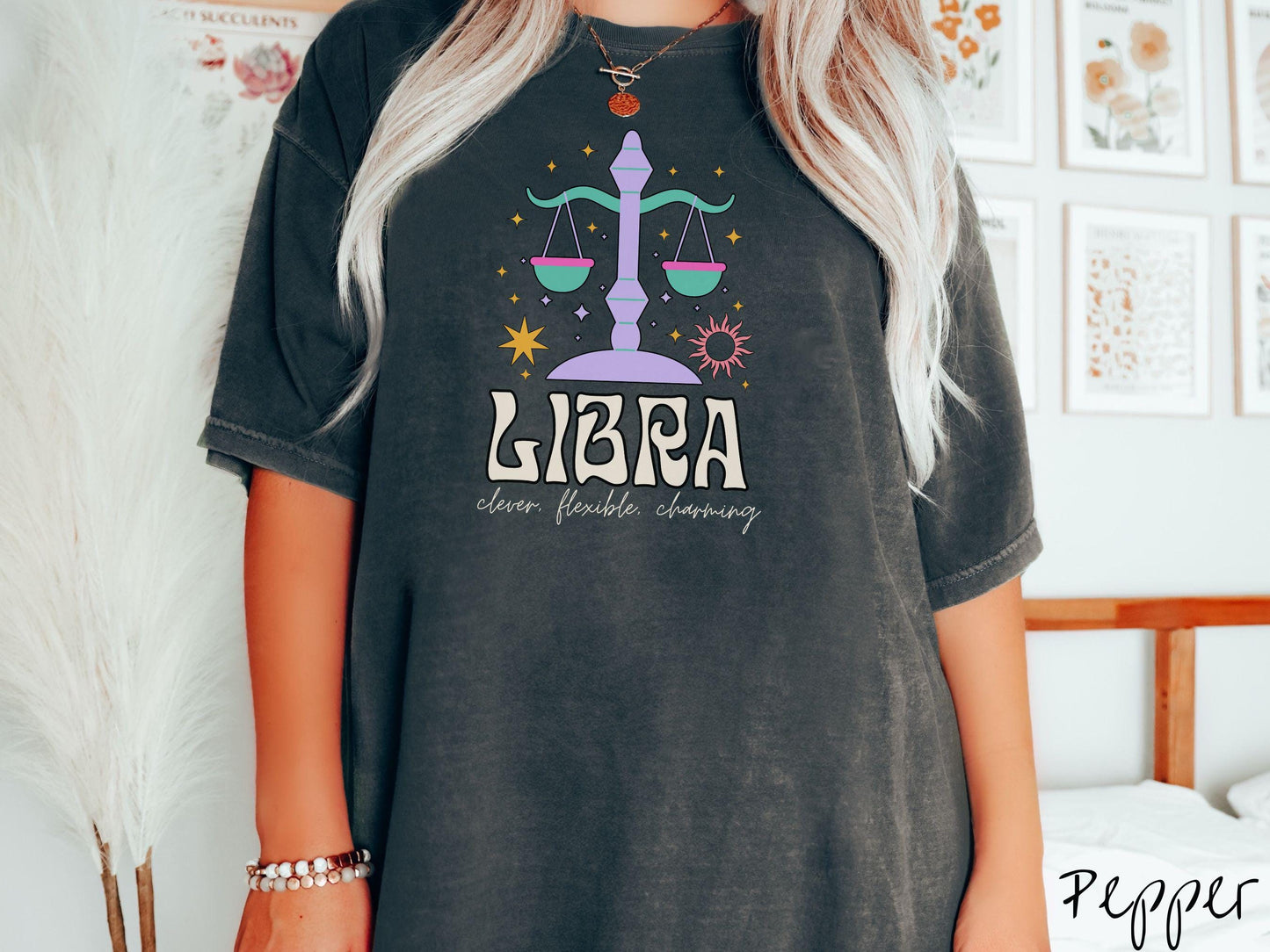 A vintage pepper colored Comfort Colors T-shirt features a large purple and green weight scale surrounded by stars and the sun with the text Libra in large bold font and below that are the words clever, flexible, charming in cursive font.