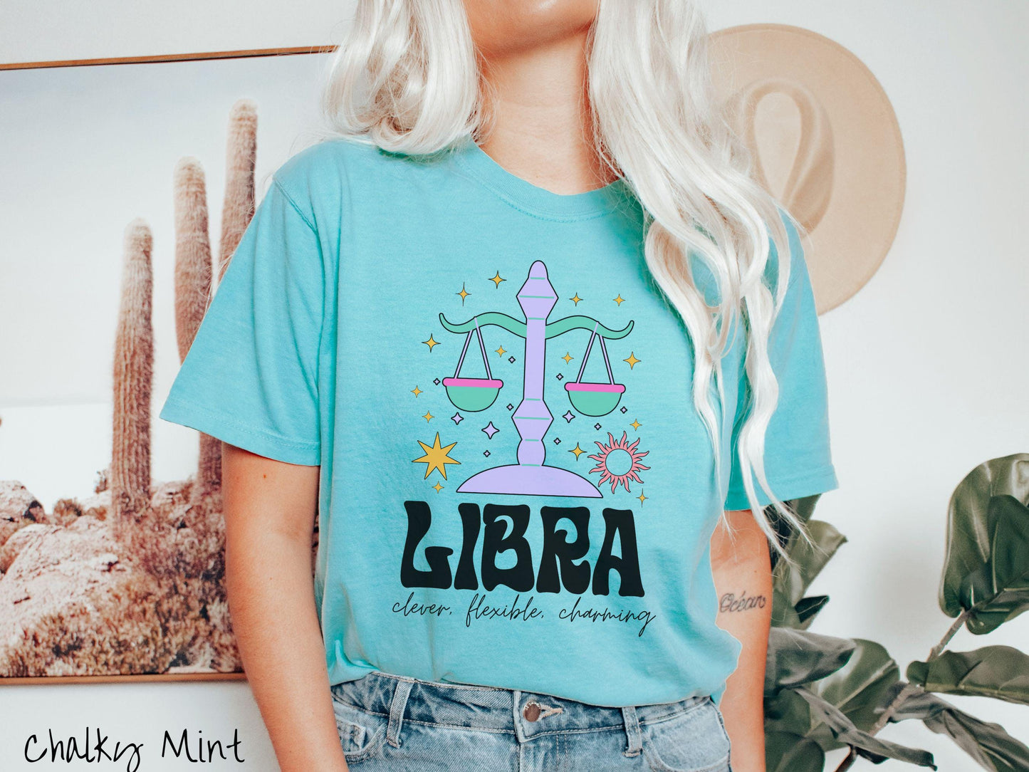 A vintage chalky mint colored Comfort Colors T-shirt features a large purple and green weight scale surrounded by stars and the sun with the text Libra in large bold font and below that are the words clever, flexible, charming in cursive font.