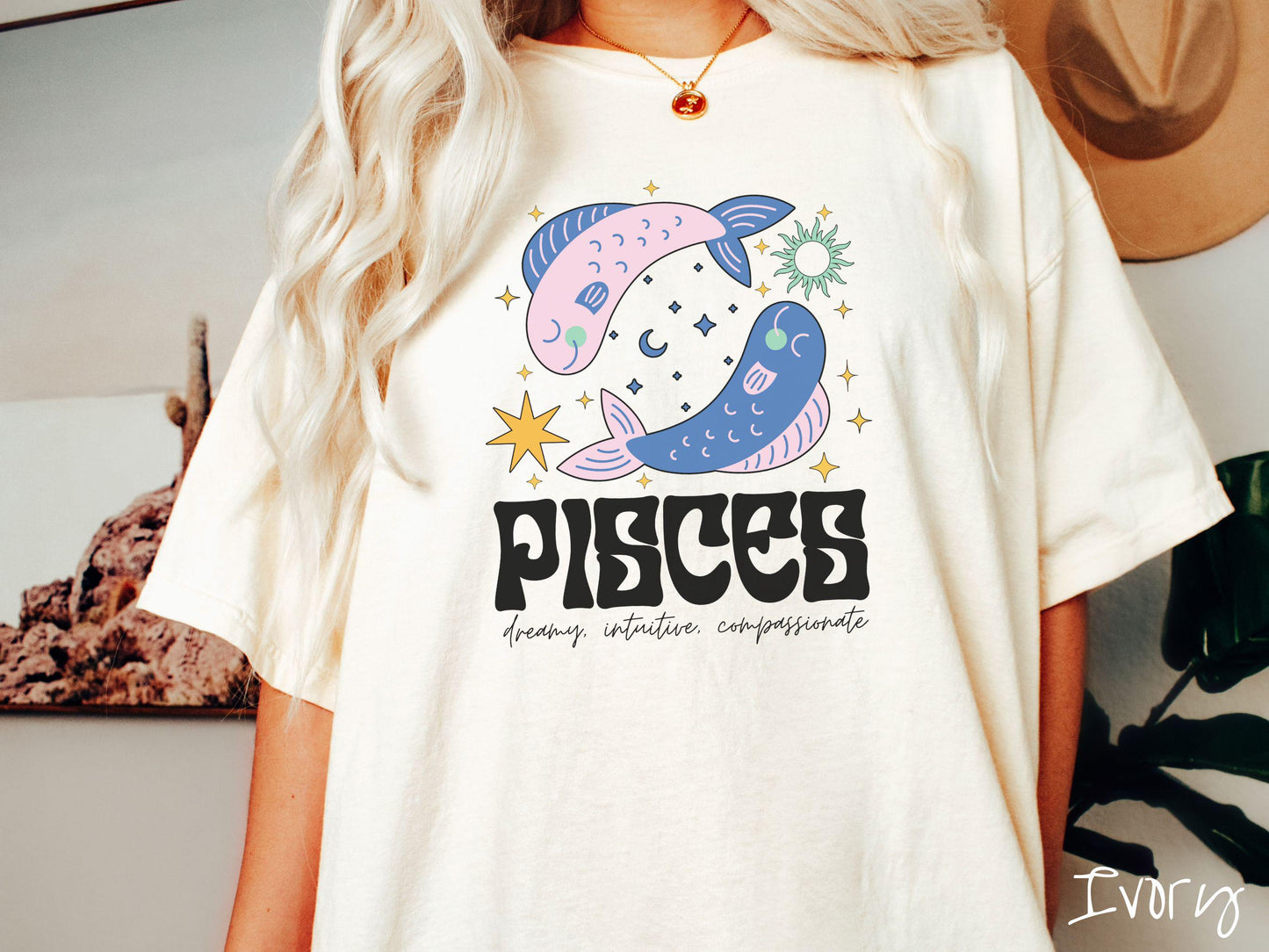 A vintage ivory colored Comfort Colors T-shirt features two large pink and blue fish surrounded by stars and the sun with the text Pisces in large bold font and below that are the words dreamy, intuitive, compassionate in cursive font.