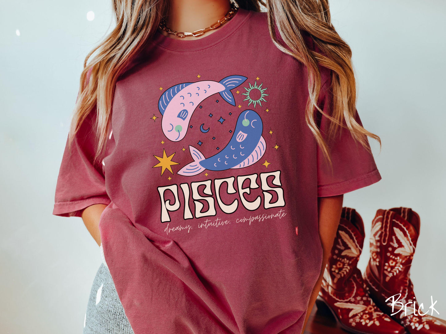 A vintage brick colored Comfort Colors T-shirt features two large pink and blue fish surrounded by stars and the sun with the text Pisces in large bold font and below that are the words dreamy, intuitive, compassionate in cursive font.