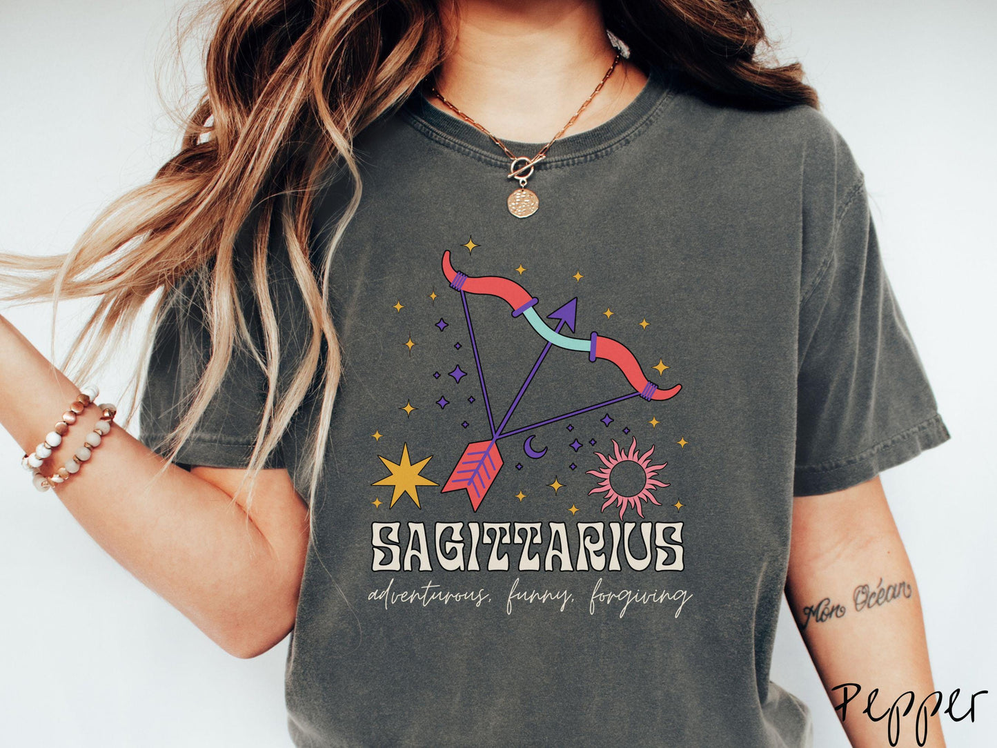 A vintage pepper colored Comfort Colors T-shirt features a large red and purple bow and arrow surrounded by stars and the sun with the text Sagittarius in large bold font and below that are the words adventurous, funny, forgiving in cursive font.
