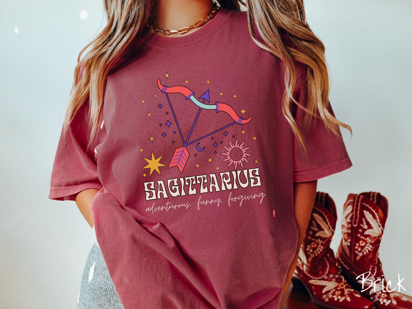 A vintage brick colored Comfort Colors T-shirt features a large red and purple bow and arrow surrounded by stars and the sun with the text Sagittarius in large bold font and below that are the words adventurous, funny, forgiving in cursive font.