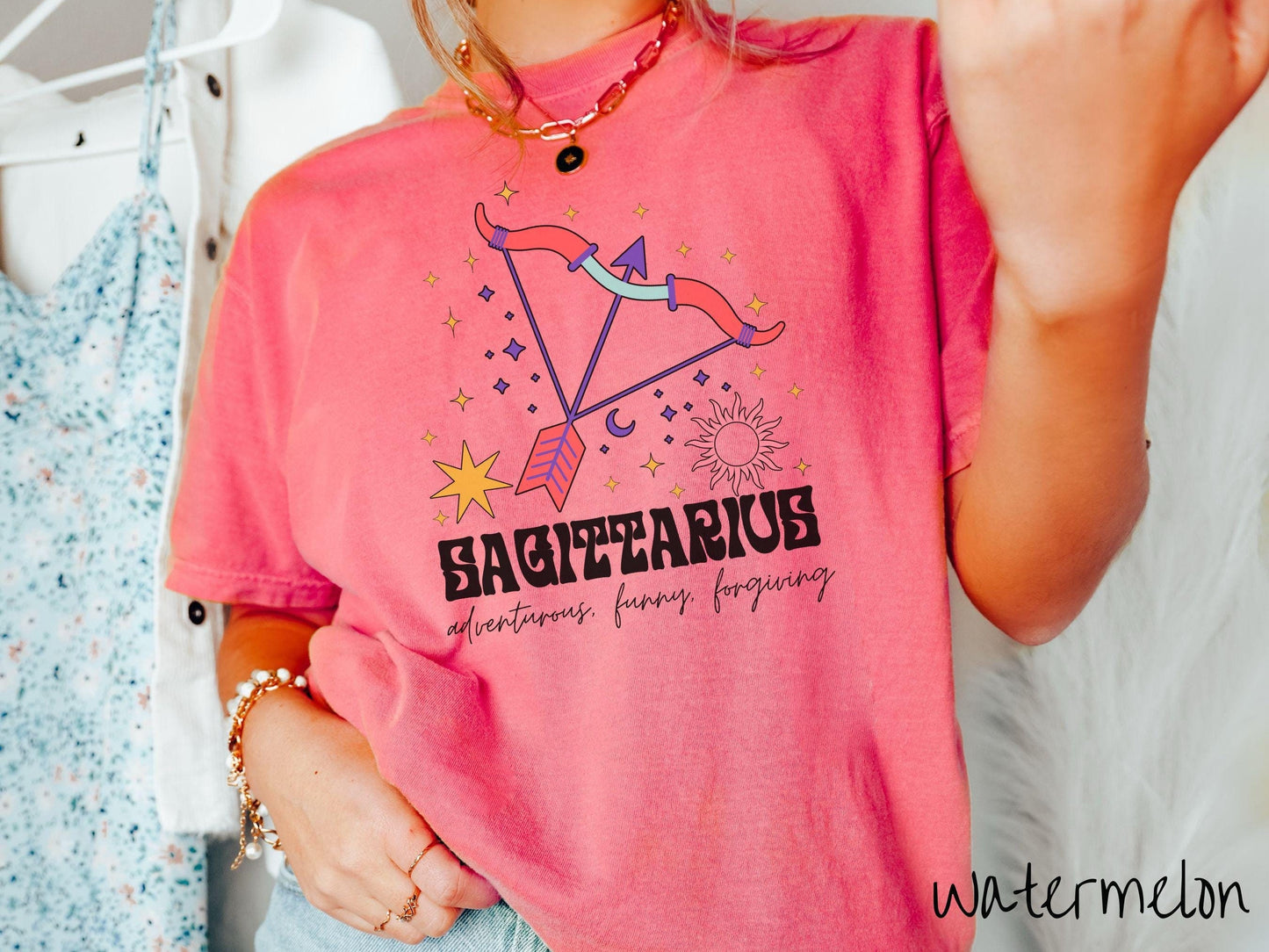 A vintage watermelon colored Comfort Colors T-shirt features a large red and purple bow and arrow surrounded by stars and the sun with the text Sagittarius in large bold font and below that are the words adventurous, funny, forgiving in cursive font.