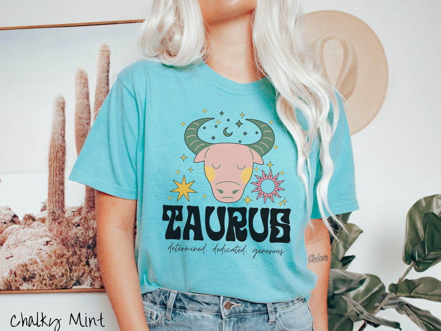 A vintage chalky mint colored Comfort Colors T-shirt features a large pink and green bull surrounded by stars and the sun with the text Taurus in large bold font and below that are the words determined, dedicated, generous in cursive font.