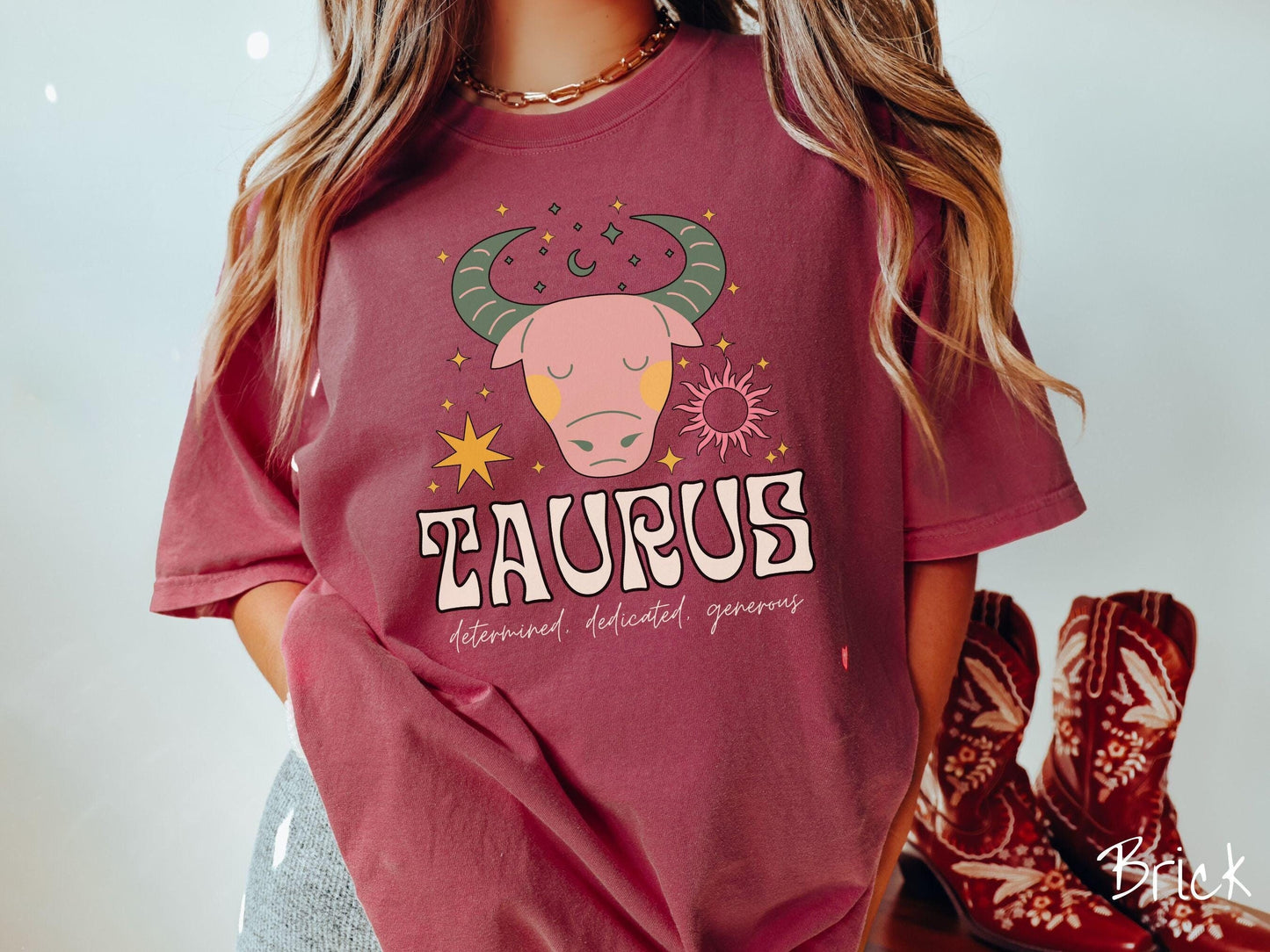 A vintage brick colored Comfort Colors T-shirt features a large pink and green bull surrounded by stars and the sun with the text Taurus in large bold font and below that are the words determined, dedicated, generous in cursive font.