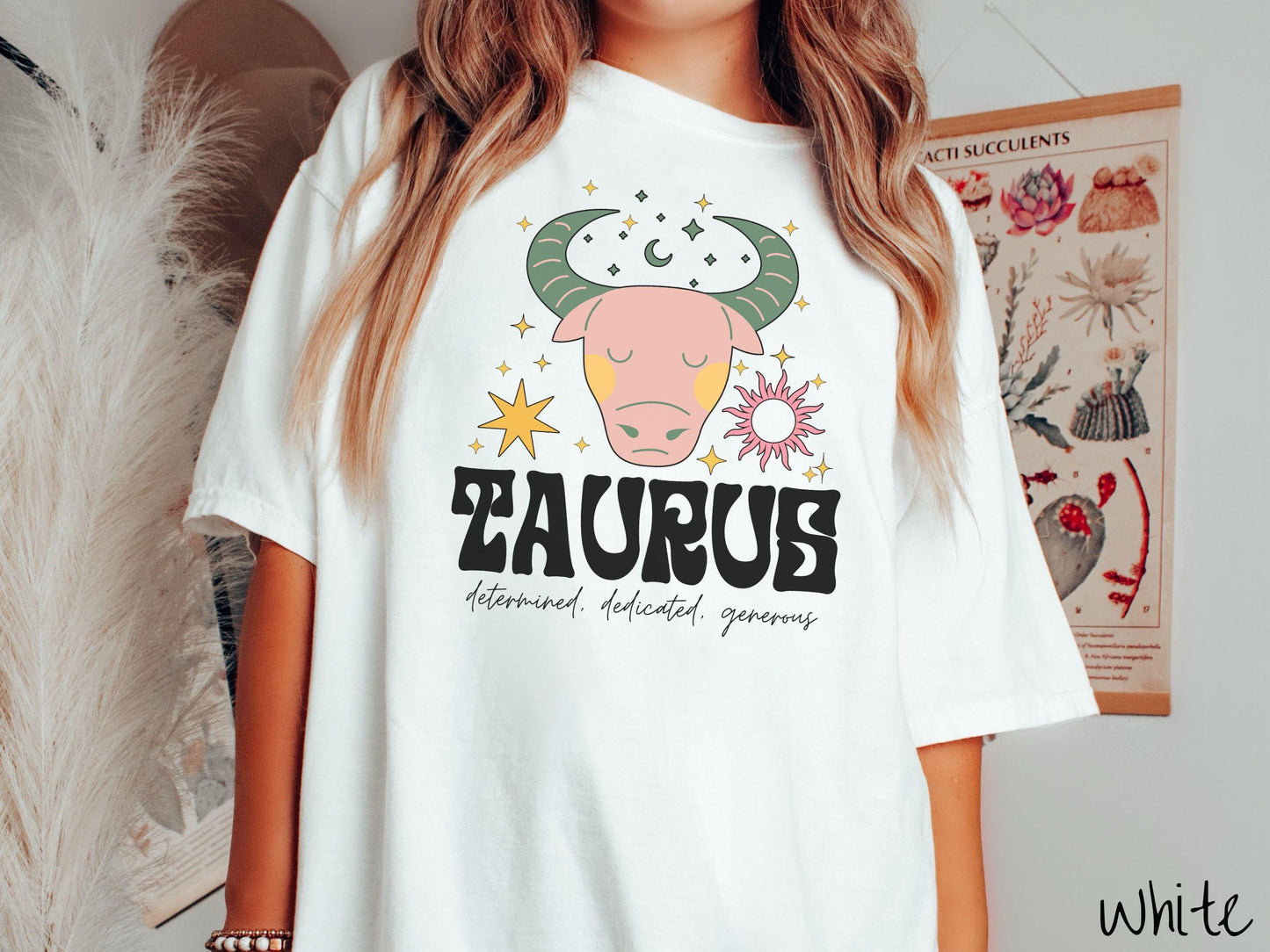 A vintage white colored Comfort Colors T-shirt features a large pink and green bull surrounded by stars and the sun with the text Taurus in large bold font and below that are the words determined, dedicated, generous in cursive font.