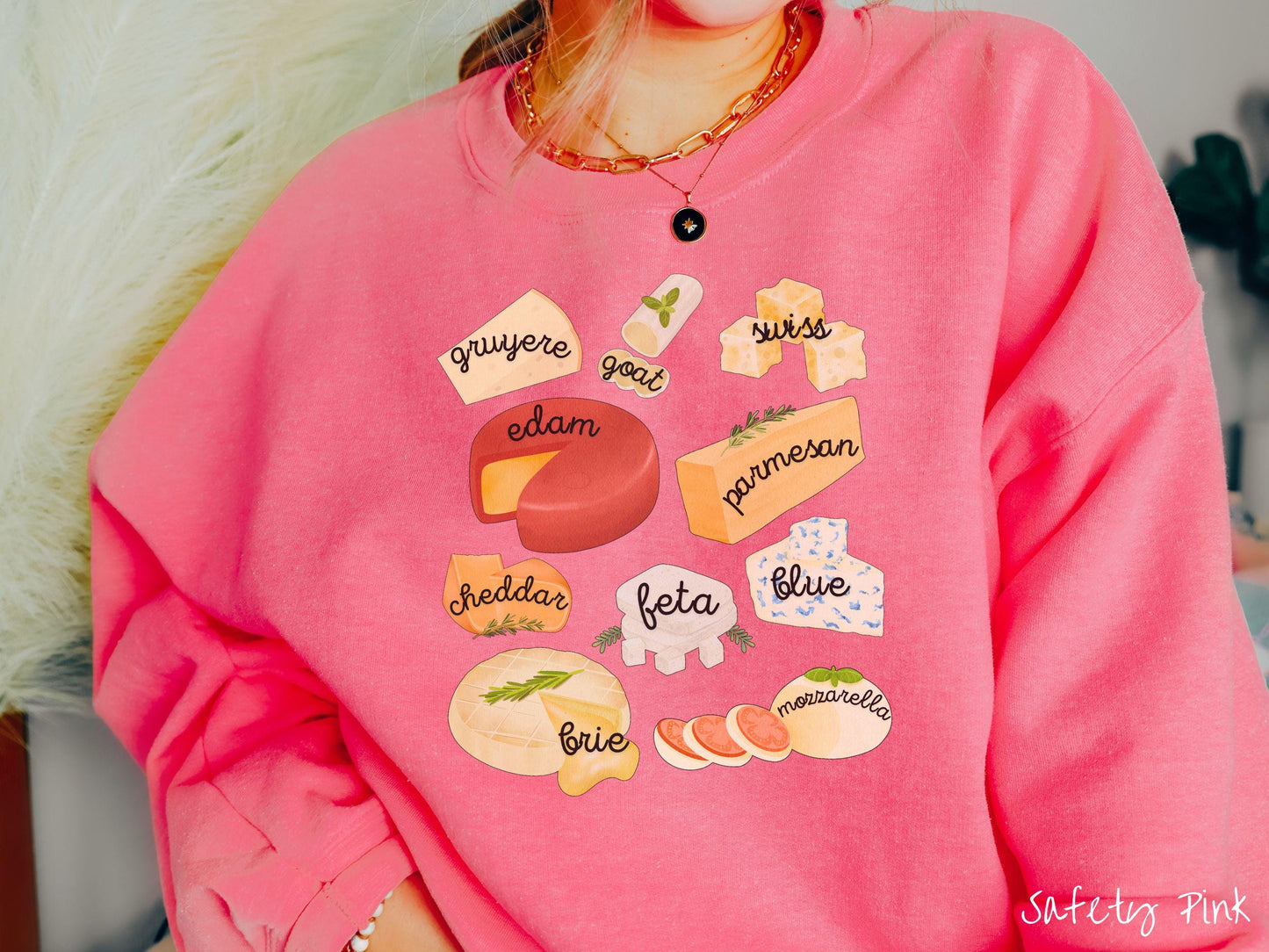 A vintage safety pink colored Gildan sweatshirt is perfect for cheese lovers with a grid pattern of various cheeses like feta, parmesan, gruyere, goat, swiss, edam, cheddar, blue, brie, and mozzarella listed.