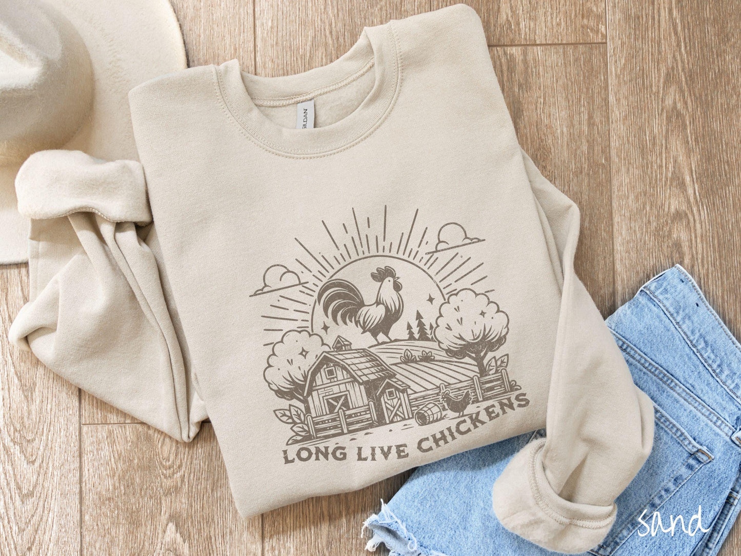 A vintage sand colored Gildan sweatshirt featuring a design of a farm with a fence, trees, and plants scattered along the property, a huge chicken in the background at the top of a pasture in front of the sun, and text Long Live Chickens.