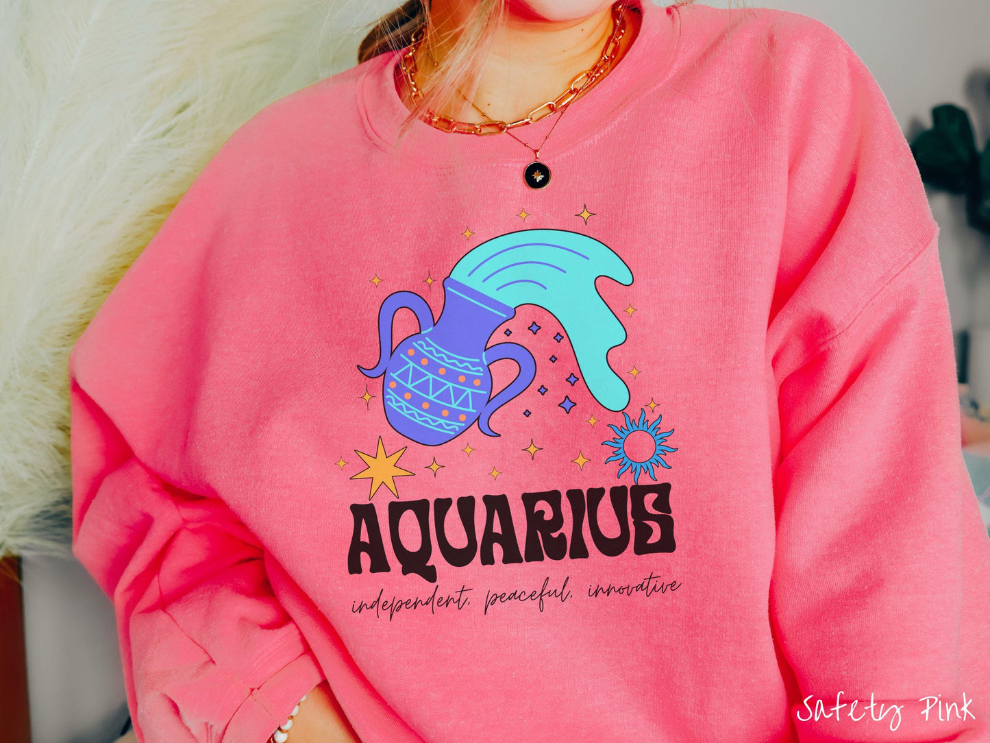 This safety pink colored vintage Comfort Colors sweatshirt features a large purple urn with blue water flowing out of it, surrounded by stars, with the text Aquarius and the words independent, peaceful, innovative.