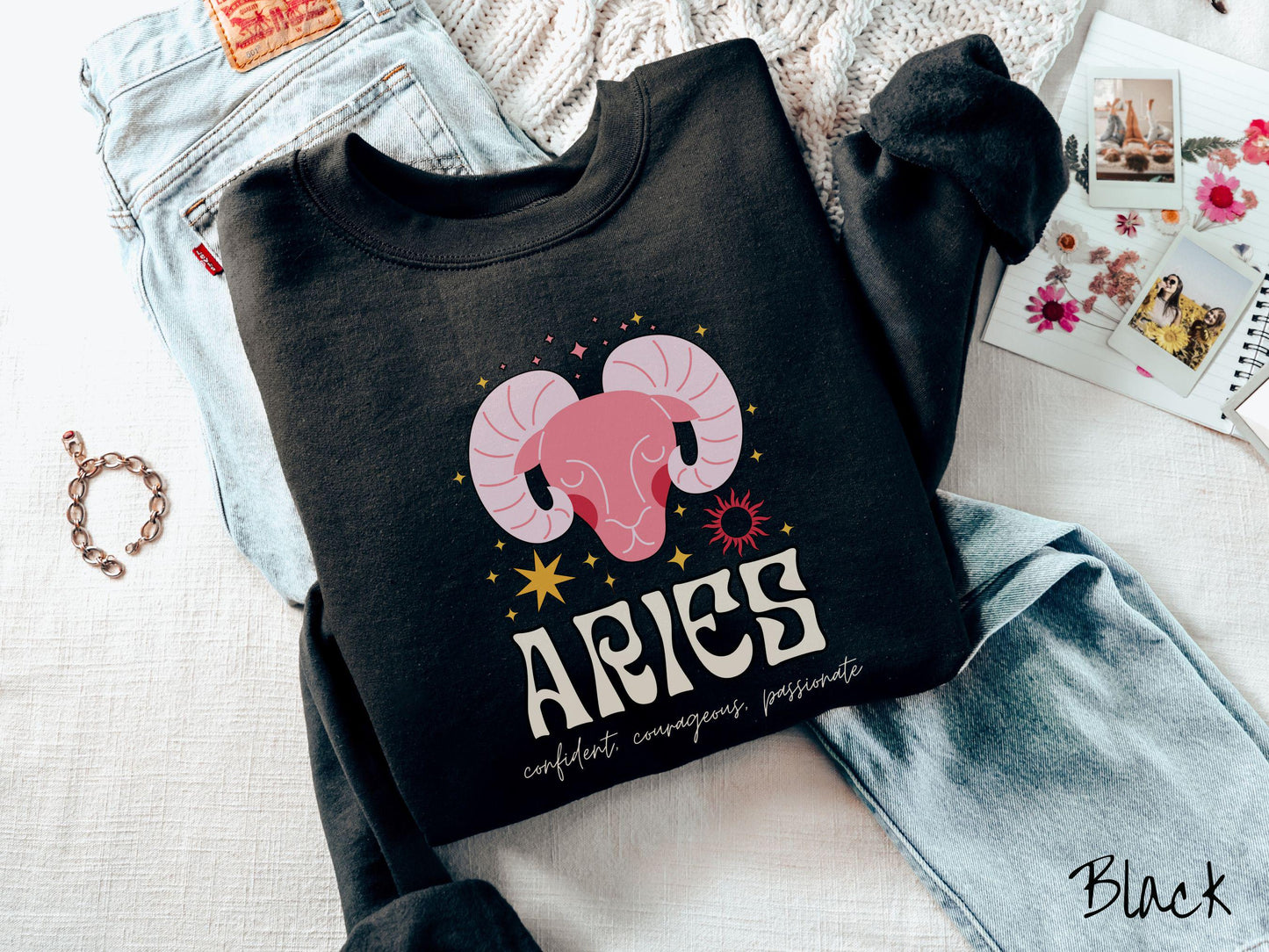 A vintage black colored Comfort Colors sweatshirt features a large red ram head surrounded by stars and the sun, with the text Aries in large bold font and below that are the words confident, courageous, passionate in cursive font.