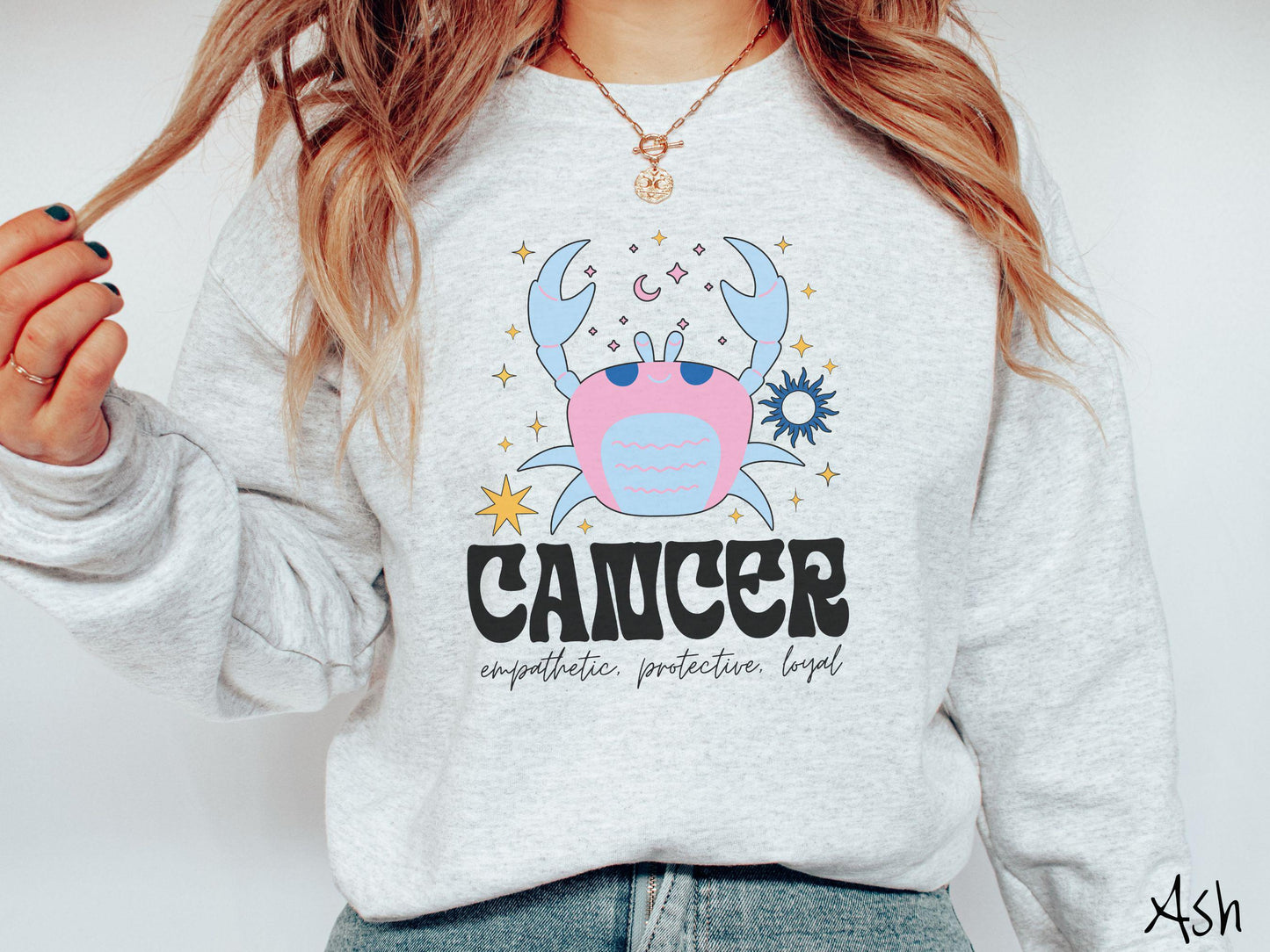 A vintage ash colored Comfort Colors sweatshirt features a large pink and blue crab, surrounded by stars, with the text Cancer and the words empathetic, protective, loyal.