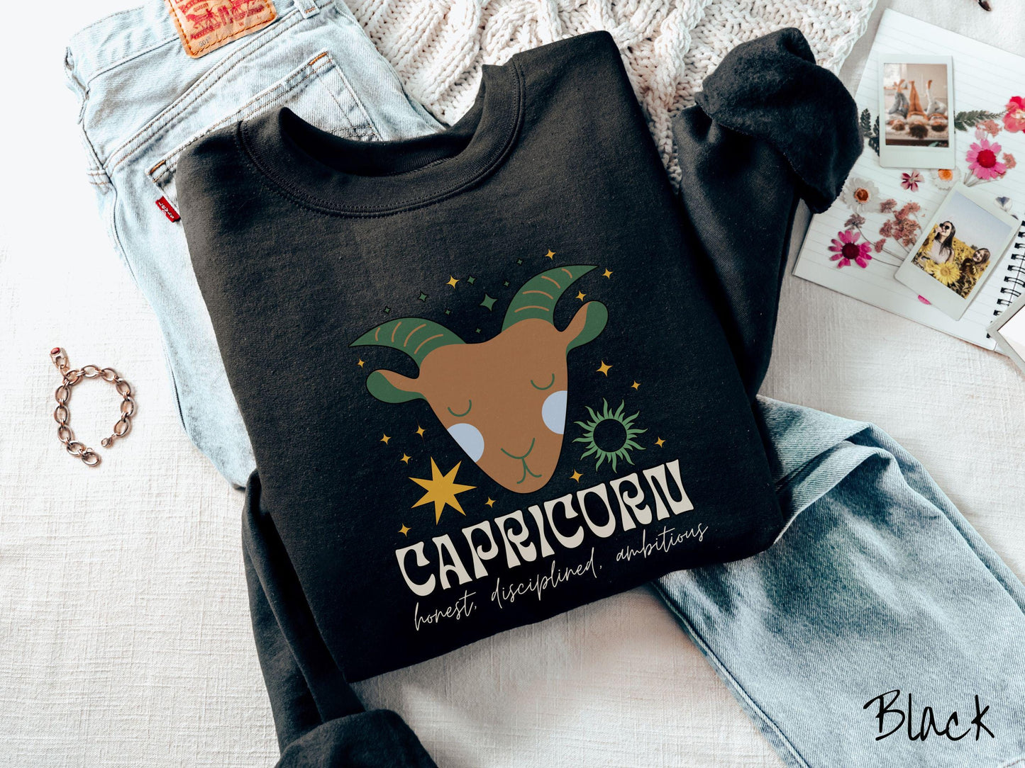 A vintage black colored Gildan sweatshirt features a large brown and green goat head surrounded by stars and the sun with the text Capricorn in large bold font and below that are the words honest, disciplined, ambitious in cursive font.