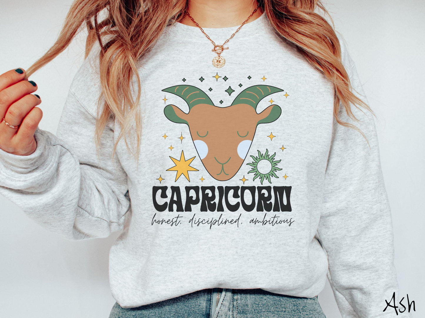 A vintage ash colored Gildan sweatshirt features a large brown and green goat head surrounded by stars and the sun with the text Capricorn in large bold font and below that are the words honest, disciplined, ambitious in cursive font.