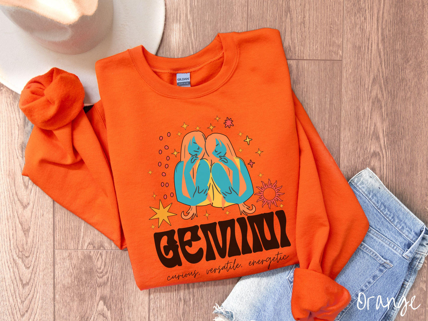 A vintage orange colored Gildan sweatshirt features the twins Castor and Pollux in blue and orange colors surrounded by stars and sun with text Gemini in large bold font and below that are the words curious, versatile, energetic in cursive font.