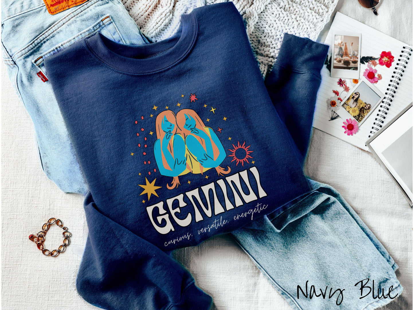 A vintage navy blue colored Gildan sweatshirt features the twins Castor and Pollux in blue and orange colors surrounded by stars and sun with text Gemini in large bold font and below that are the words curious, versatile, energetic in cursive font.