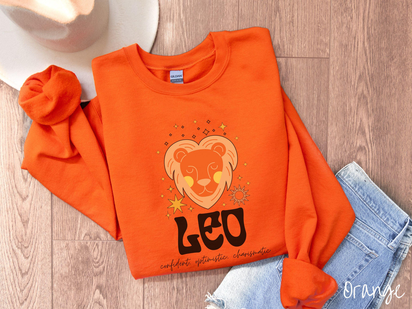 A vintage orange colored Gildan sweatshirt features a large orange and yellow lion surrounded by stars and the sun with the text Leo in large bold font and below that are the words confident, optimistic, charismatic in cursive font.