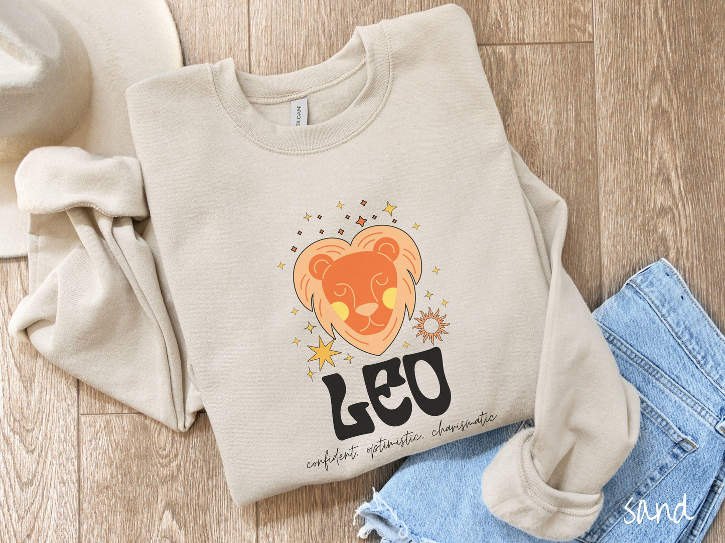 A vintage sand colored Gildan sweatshirt features a large orange and yellow lion surrounded by stars and the sun with the text Leo in large bold font and below that are the words confident, optimistic, charismatic in cursive font.