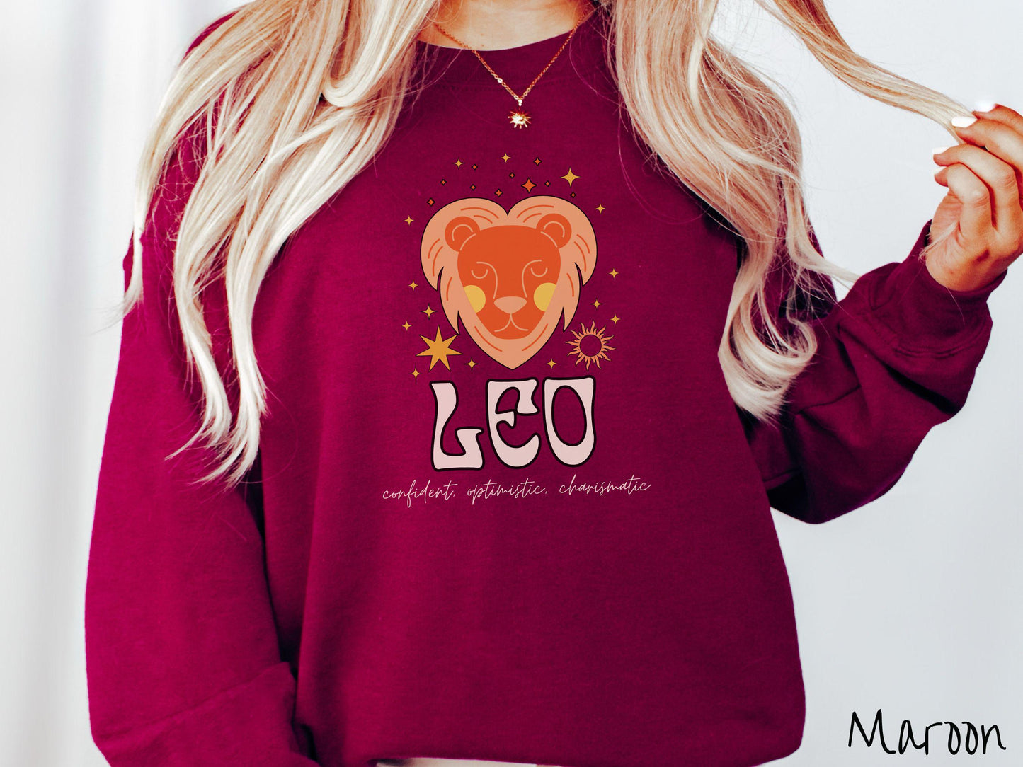 A vintage maroon colored Gildan sweatshirt features a large orange and yellow lion surrounded by stars and the sun with the text Leo in large bold font and below that are the words confident, optimistic, charismatic in cursive font.