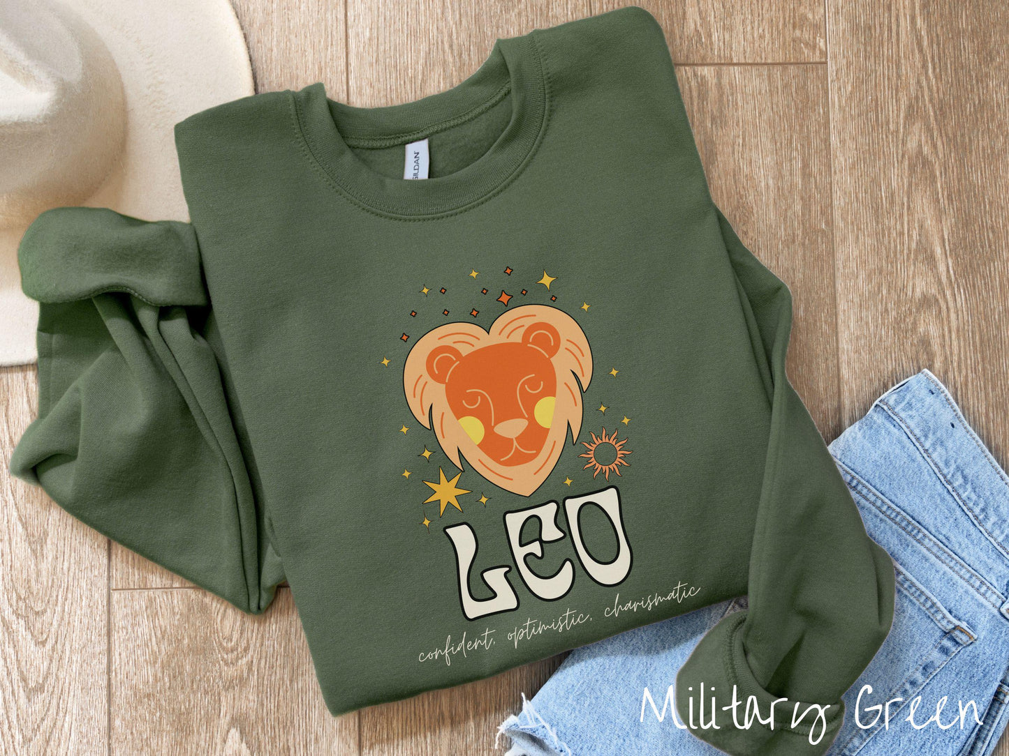 A vintage military green colored Gildan sweatshirt features a large orange and yellow lion surrounded by stars and the sun with the text Leo in large bold font and below that are the words confident, optimistic, charismatic in cursive font.