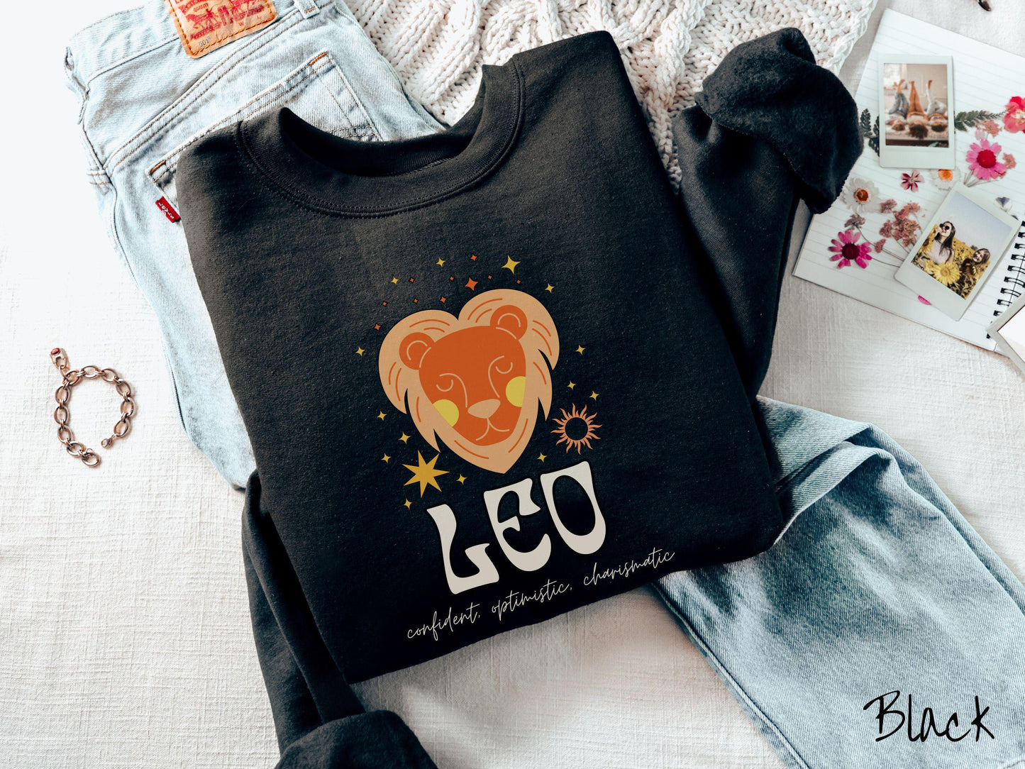A vintage black colored Gildan sweatshirt features a large orange and yellow lion surrounded by stars and the sun with the text Leo in large bold font and below that are the words confident, optimistic, charismatic in cursive font.
