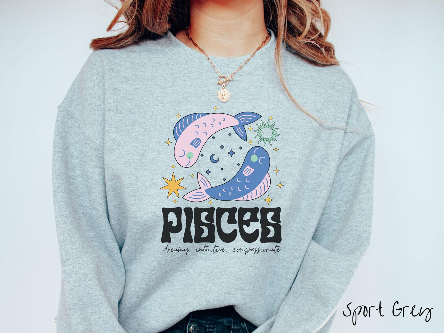 A vintage sport grey colored Gildan sweatshirt features two large pink and blue fish surrounded by stars and the sun with the text Pisces in large bold font and below that are the words dreamy, intuitive, compassionate in cursive font.