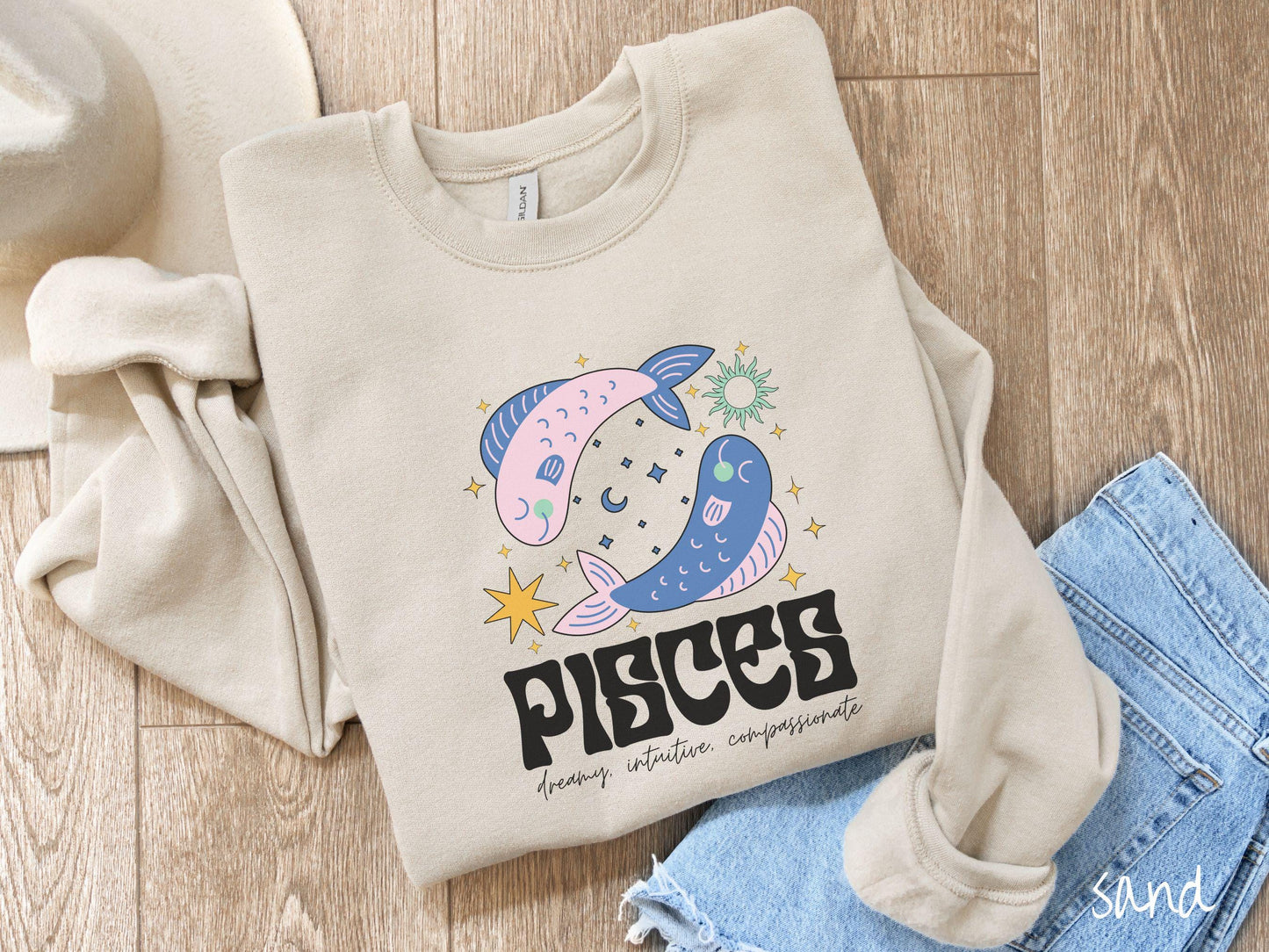 A vintage sand colored Gildan sweatshirt features two large pink and blue fish surrounded by stars and the sun with the text Pisces in large bold font and below that are the words dreamy, intuitive, compassionate in cursive font.