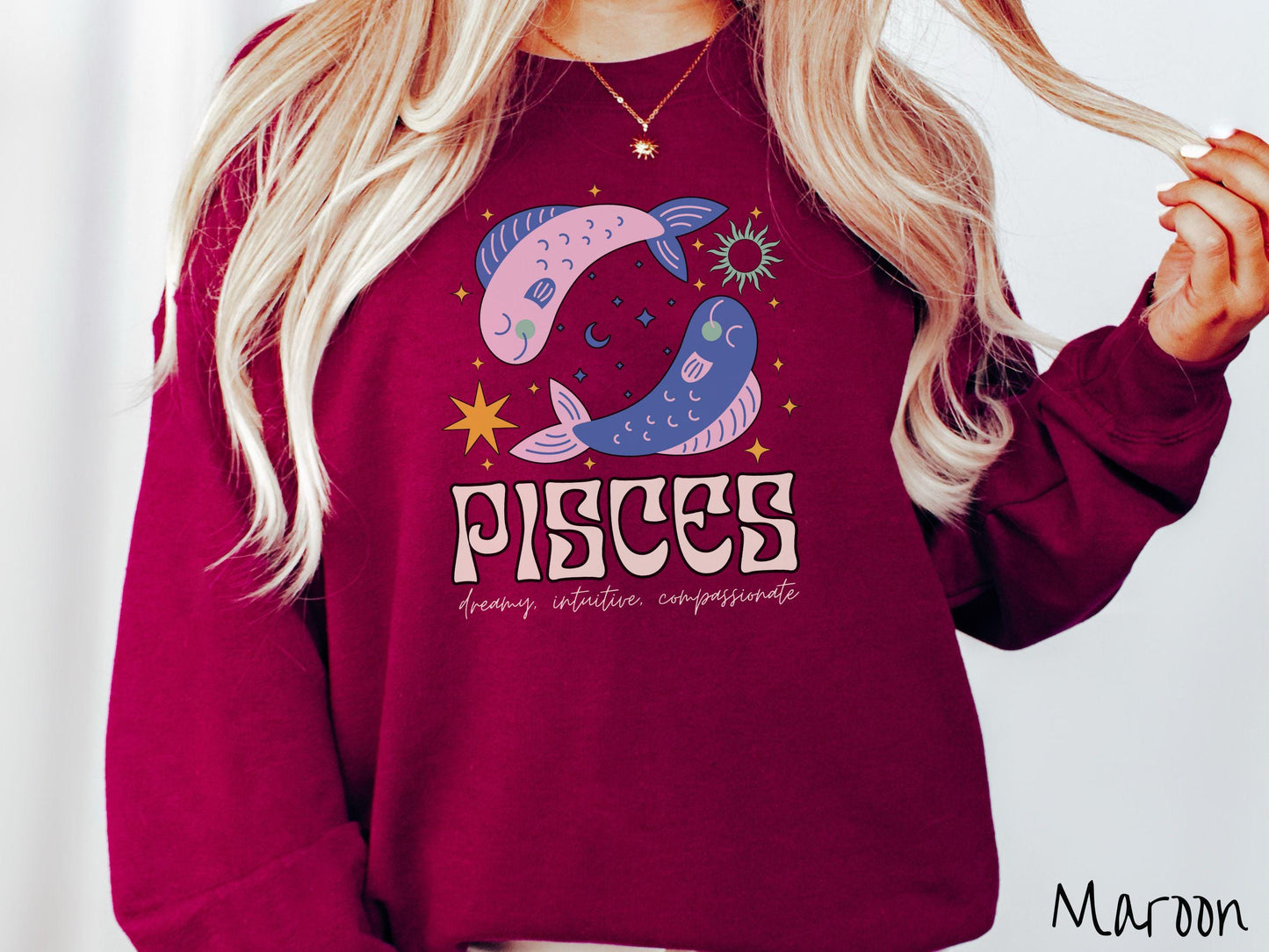 A vintage maroon colored Gildan sweatshirt features two large pink and blue fish surrounded by stars and the sun with the text Pisces in large bold font and below that are the words dreamy, intuitive, compassionate in cursive font.