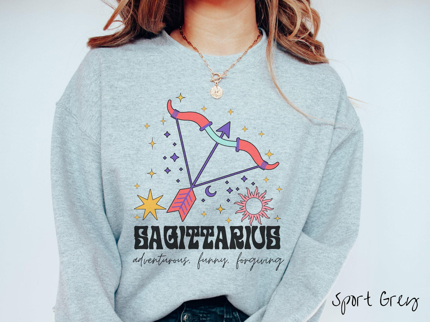 A vintage sport grey colored Gildan sweatshirt features a large red and purple bow and arrow surrounded by stars and the sun with the text Sagittarius in large bold font and below that are the words adventurous, funny, forgiving in cursive font.
