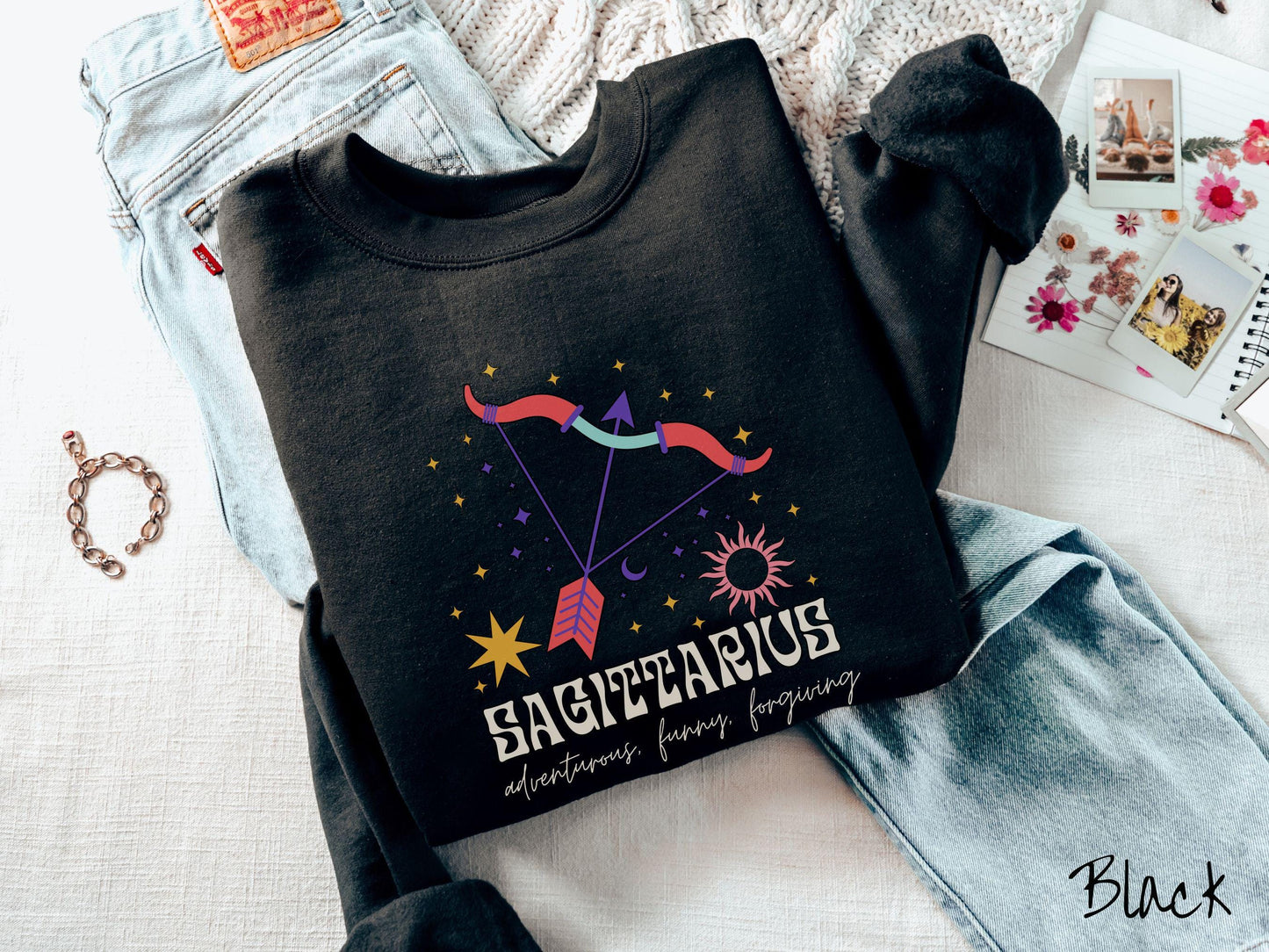 A vintage black colored Gildan sweatshirt features a large red and purple bow and arrow surrounded by stars and the sun with the text Sagittarius in large bold font and below that are the words adventurous, funny, forgiving in cursive font.
