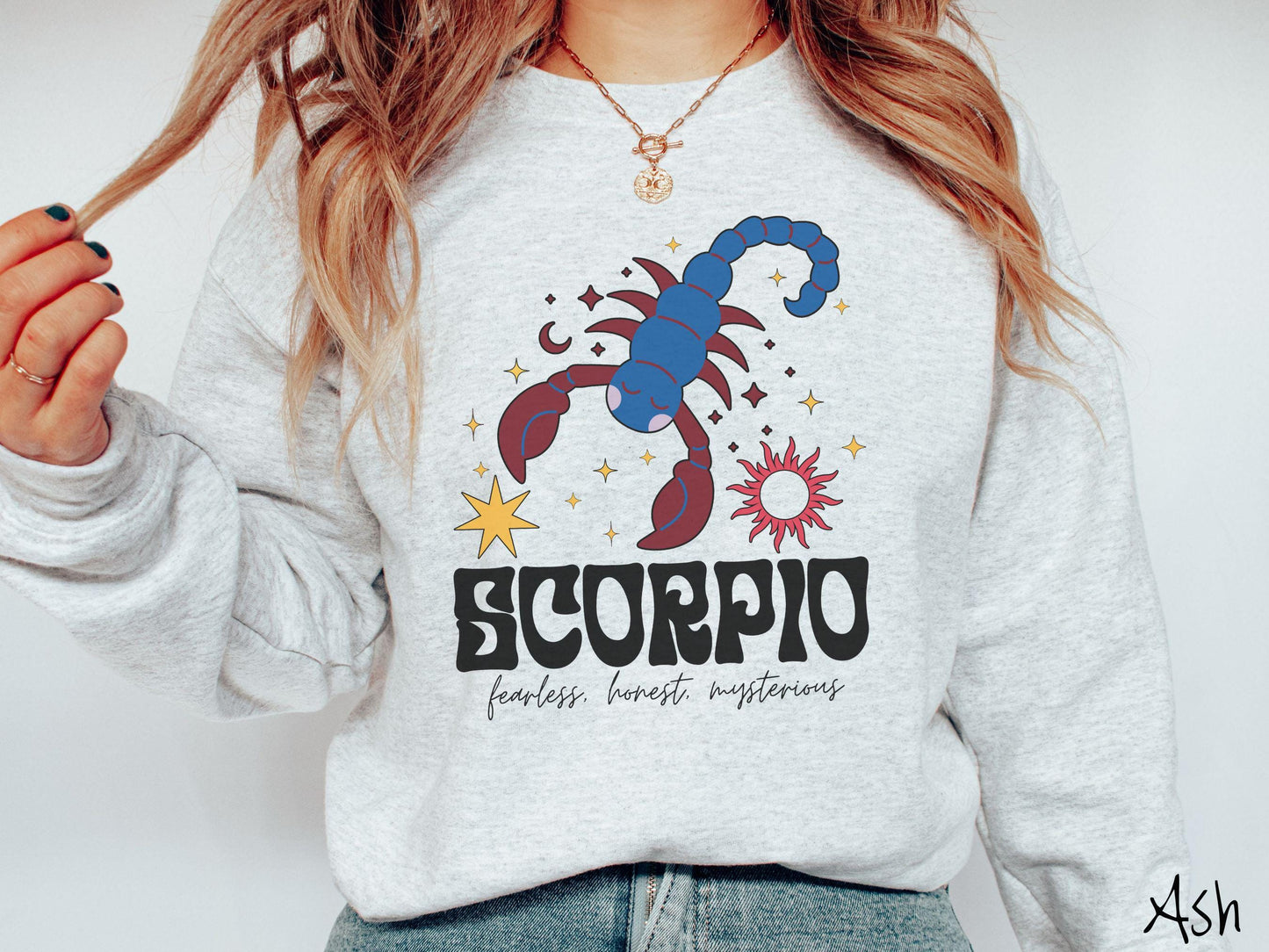 A vintage ash colored Gildan sweatshirt features a large red and blue scorpion surrounded by stars and the sun with the text Scorpio in large bold font and below that are the words fearless, honest, mysterious in cursive font.