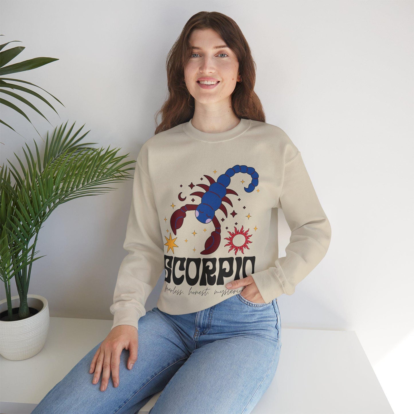 A vintage sand colored Gildan sweatshirt features a large red and blue scorpion surrounded by stars and the sun with the text Scorpio in large bold font and below that are the words fearless, honest, mysterious in cursive font.