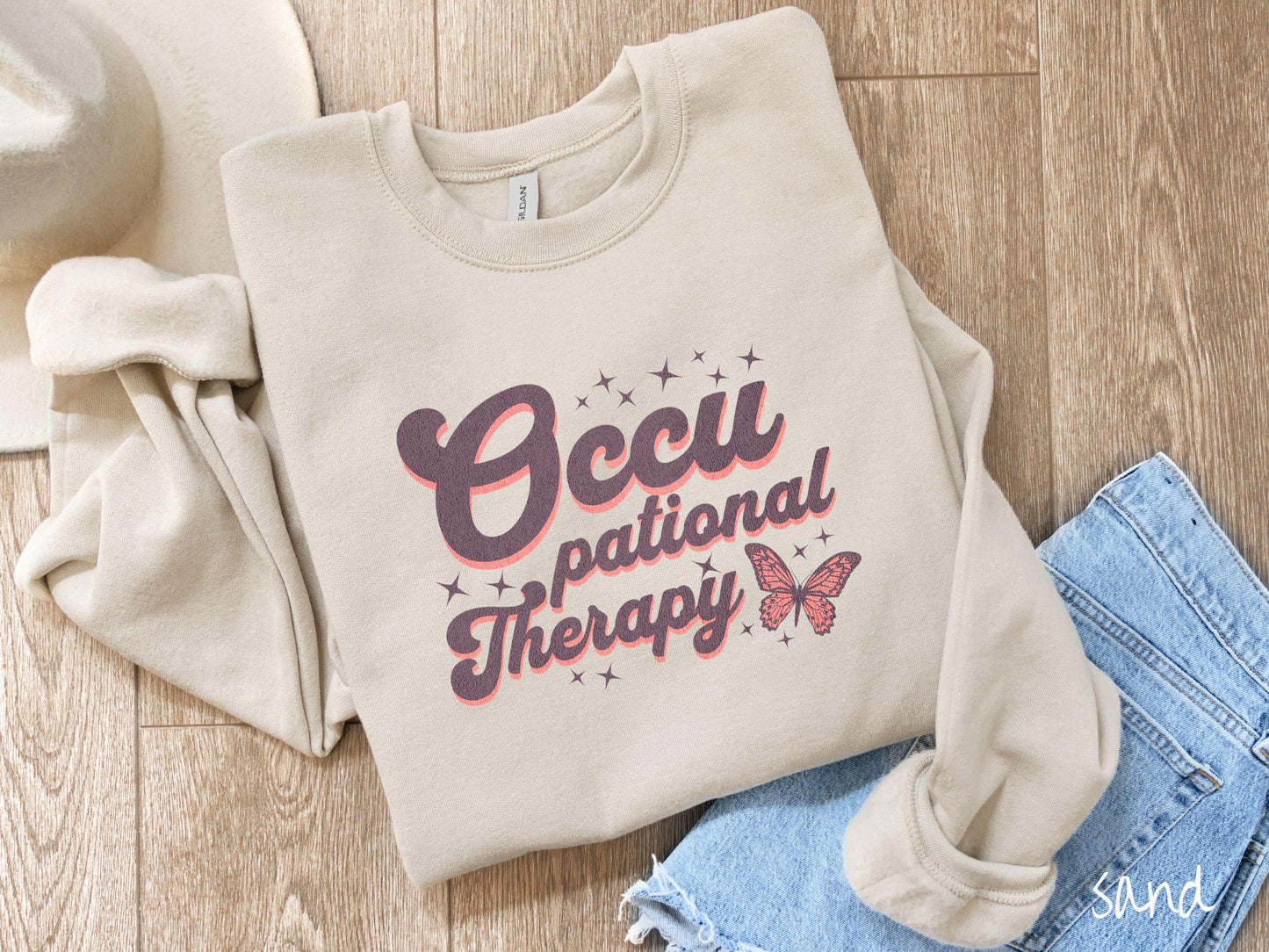 A vintage, sand colored Gildan sweatshirt featuring the words Occupational Therapy across the front, this unique design showcases a stunning pink and dark purple butterfly surrounded by delicate twinkling stars.