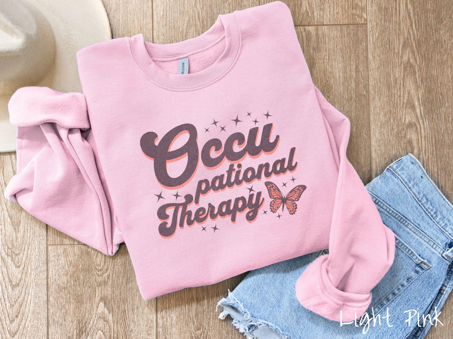 A vintage, light pink colored Gildan sweatshirt featuring the words Occupational Therapy across the front, this unique design showcases a stunning pink and dark purple butterfly surrounded by delicate twinkling stars.