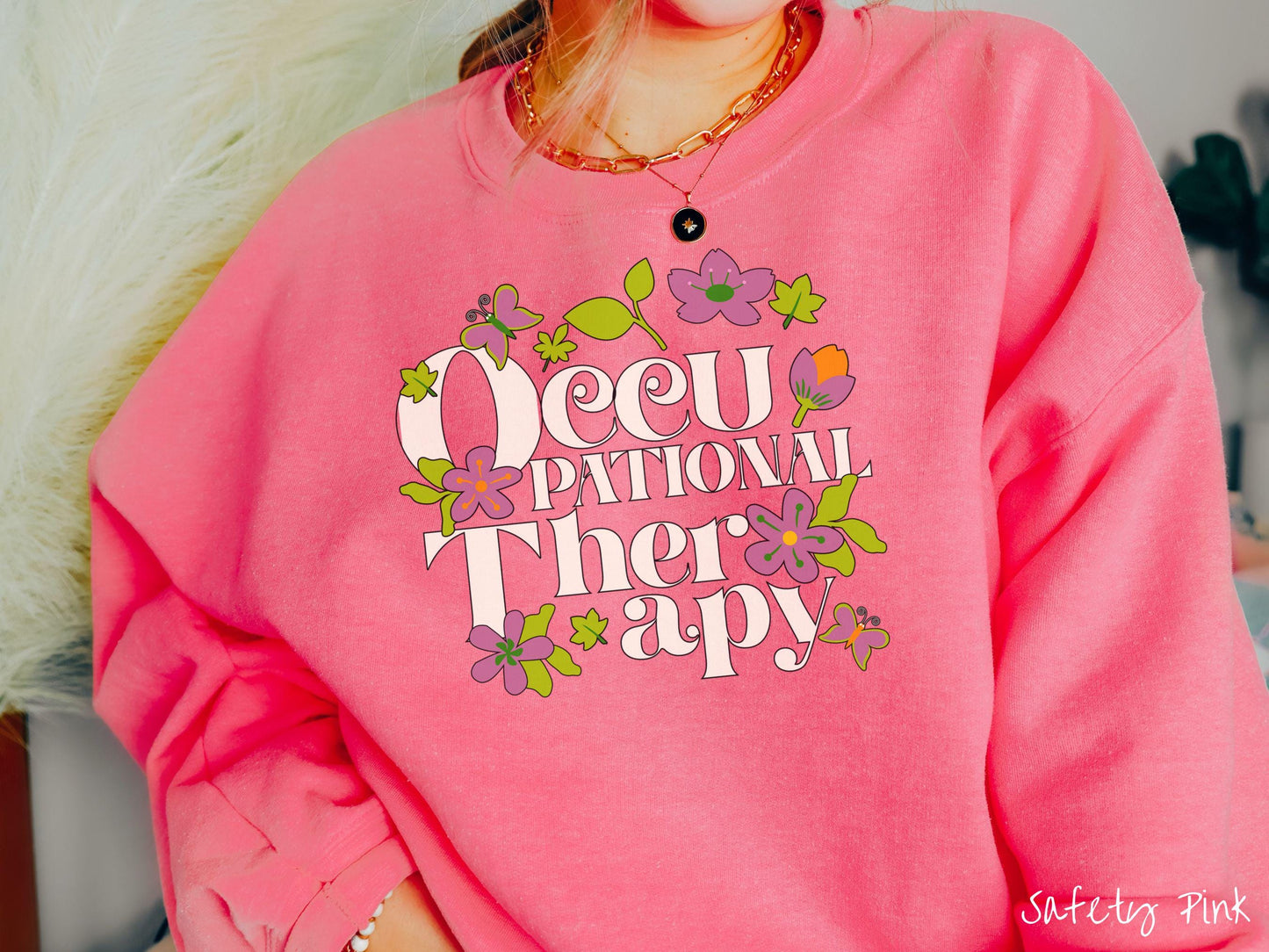 A vintage, safety pink colored Gildan sweatshirt with the text Occupational Therapy and featuring green leaves, purple and orange flowers, and colorful butterflies in the background.