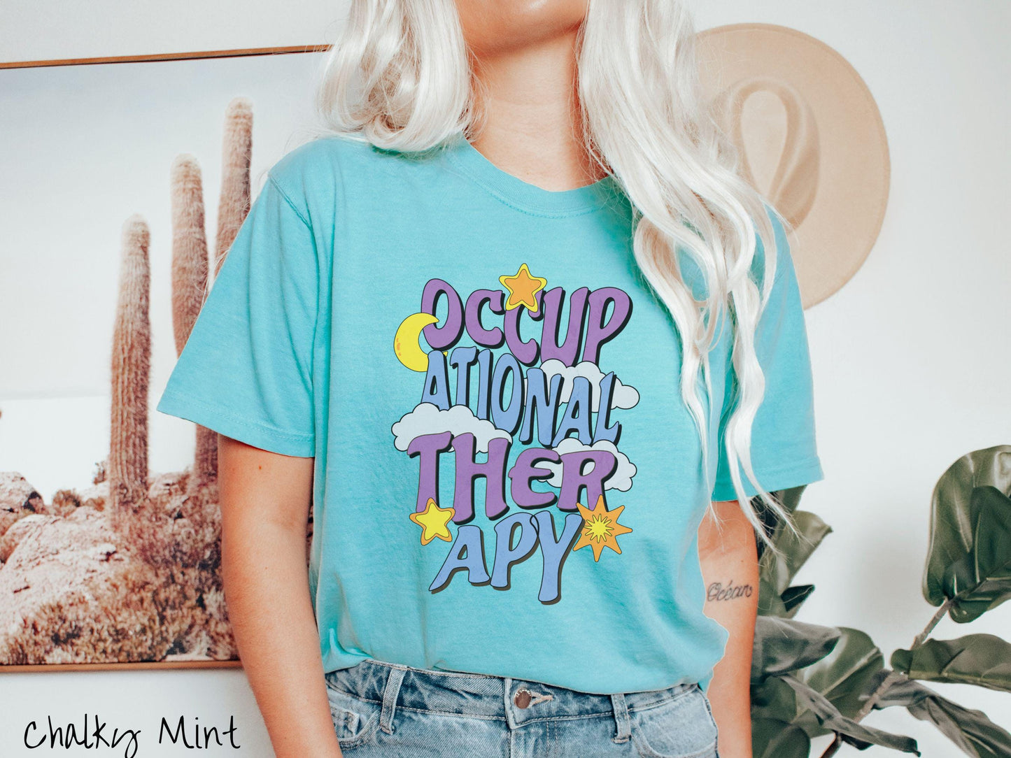 A vintage, chalky mint colored Comfort Colors t-shirt with the text Occupational Therapy vertically with clouds, the yellow moon, and bright yellow and orange stars mixed in.