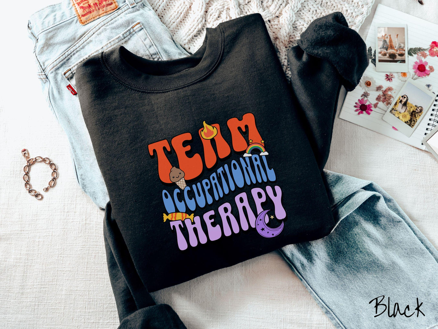 A vintage, black colored Gildan sweatshirt with the text "Team Occupational Therapy" surrounded by playful elements like a chocolate ice cream cone, a blazing flame, a colorful rainbow, a piece of candy, and a serene moon.