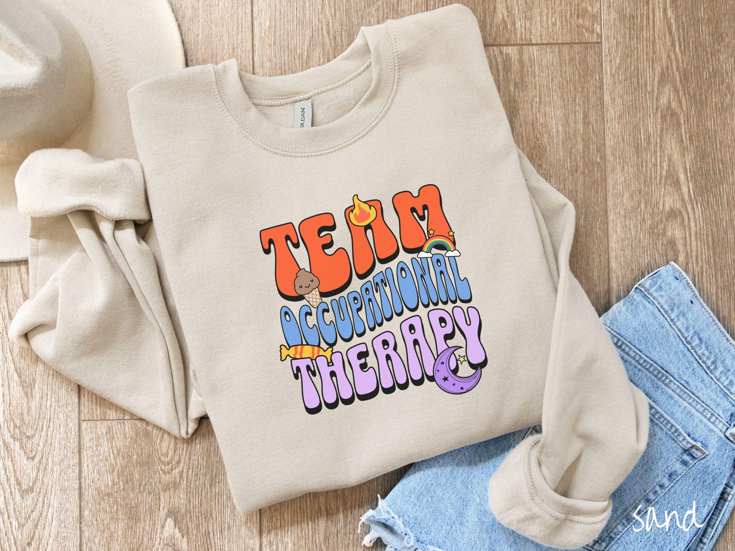 A vintage, sand colored Gildan sweatshirt with the text "Team Occupational Therapy" surrounded by playful elements like a chocolate ice cream cone, a blazing flame, a colorful rainbow, a piece of candy, and a serene moon.