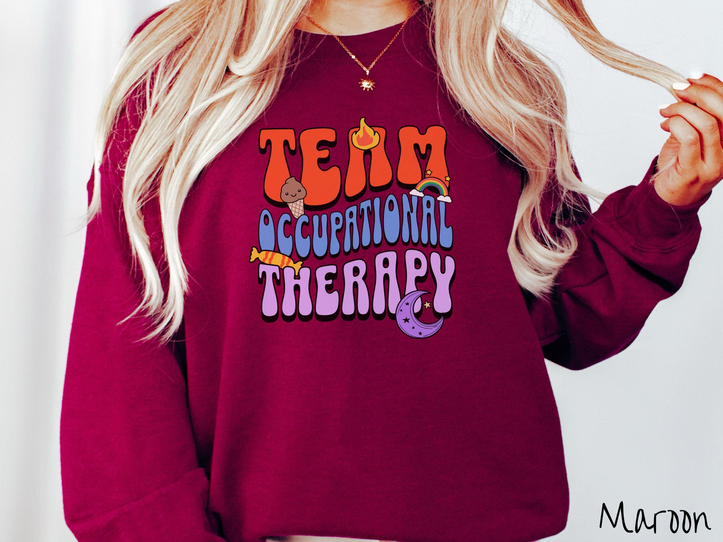A vintage, maroon colored Gildan sweatshirt with the text "Team Occupational Therapy" surrounded by playful elements like a chocolate ice cream cone, a blazing flame, a colorful rainbow, a piece of candy, and a serene moon.
