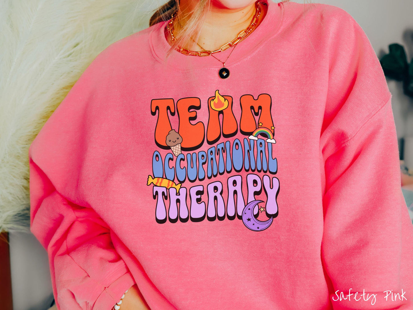 A vintage, safety pink colored Gildan sweatshirt with the text "Team Occupational Therapy" surrounded by playful elements like a chocolate ice cream cone, a blazing flame, a colorful rainbow, a piece of candy, and a serene moon.