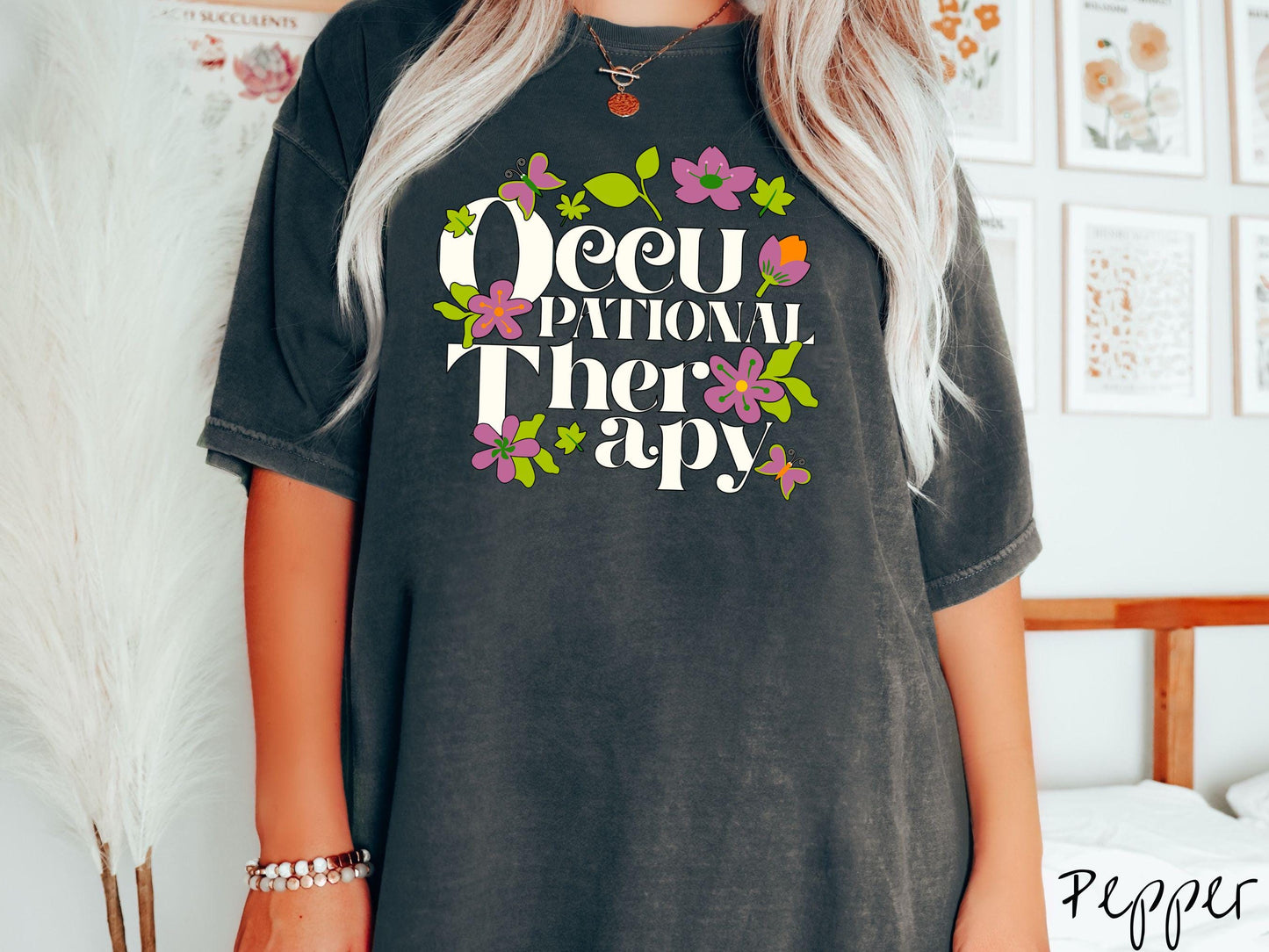 A vintage, pepper colored Comfort Colors t-shirt with the text Occupational Therapy and featuring green leaves, purple and orange flowers, and colorful butterflies in the background.