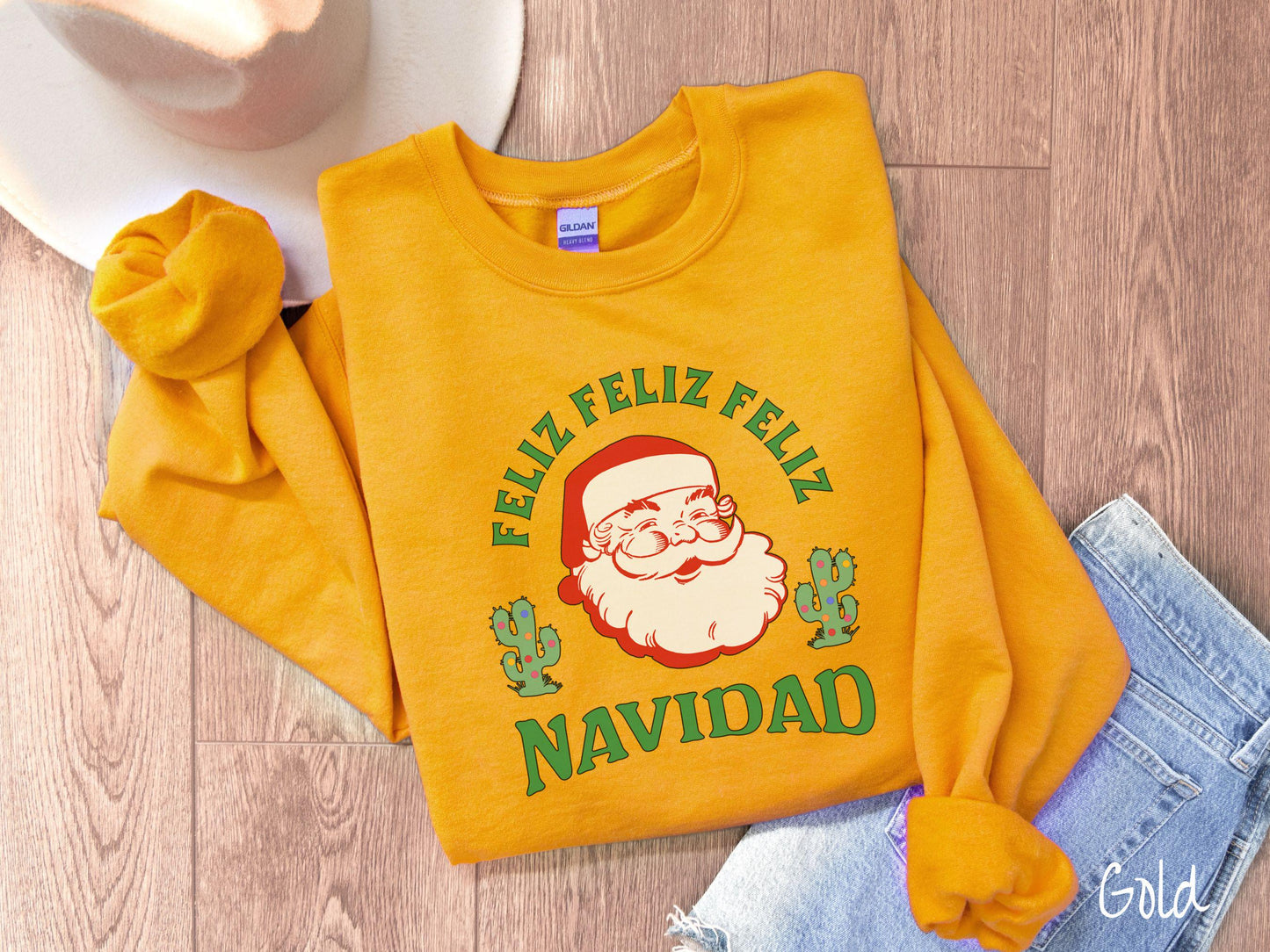 A festive gold colored Gildan sweatshirt featuring a smiling santa clause between two green cacti covered in colorful Christmas lights and the phrase, 'Feliz feliz feliz Navidad' is perfect for spreading holiday cheer.