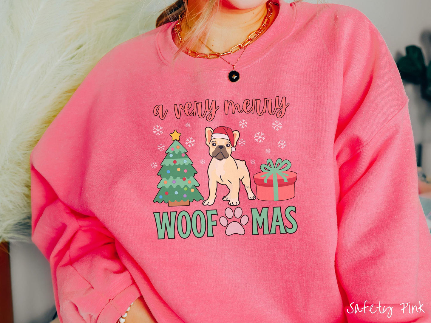 A vintage safety pink colored Gildan sweatshirt with the text 'a very merry Woofmas' in red and green font, featuring a French Bulldog wearing a red Santa hat sitting between a Christmas tree and a present with white snowflakes hanging in the air.
