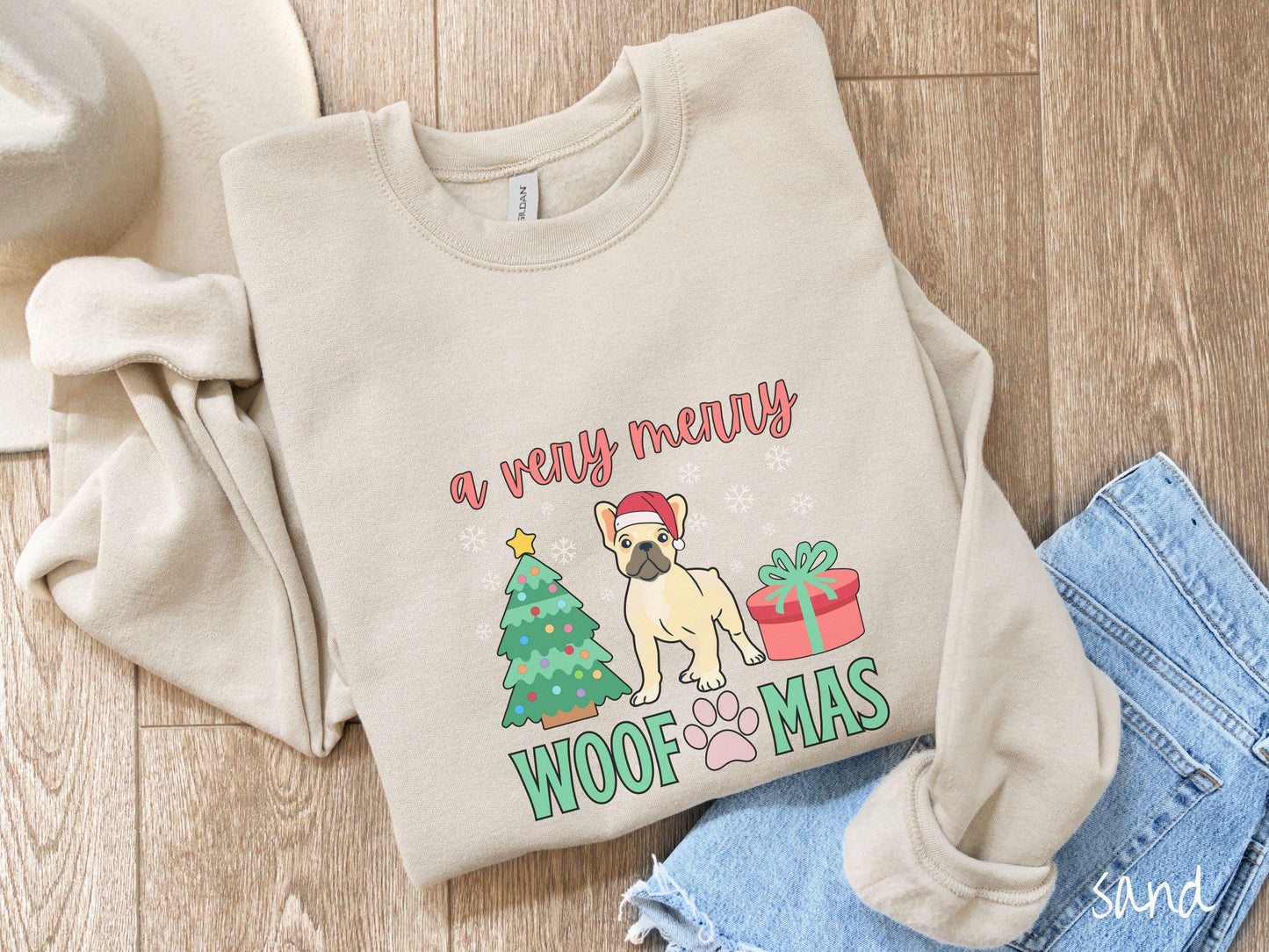 A vintage sand colored Gildan sweatshirt with the text 'a very merry Woofmas' in red and green font, featuring a French Bulldog wearing a red Santa hat sitting between a decorated Christmas tree and a present with white snowflakes hanging in the air.