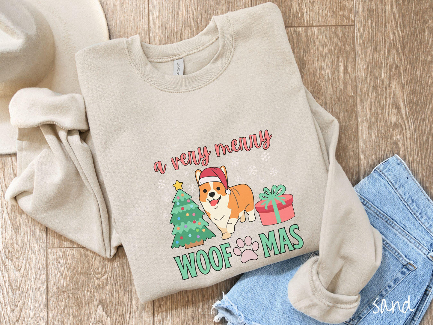 A vintage sand colored Gildan sweatshirt with the text 'a very merry Woofmas' in red and green font, featuring an orange corgi wearing a red Santa hat sitting between a Christmas tree and a present with white snowflakes hanging in the air.