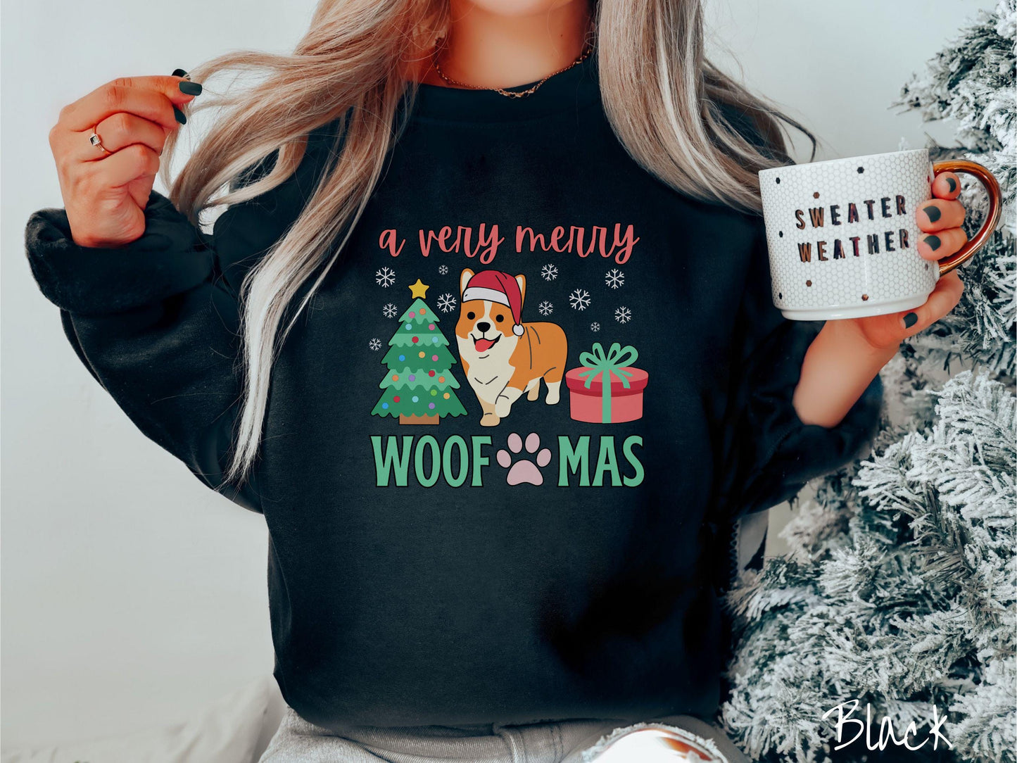 A vintage black colored Gildan sweatshirt with the text 'a very merry Woofmas' in red and green font, featuring an orange corgi wearing a red Santa hat sitting between a Christmas tree and a present with white snowflakes hanging in the air.