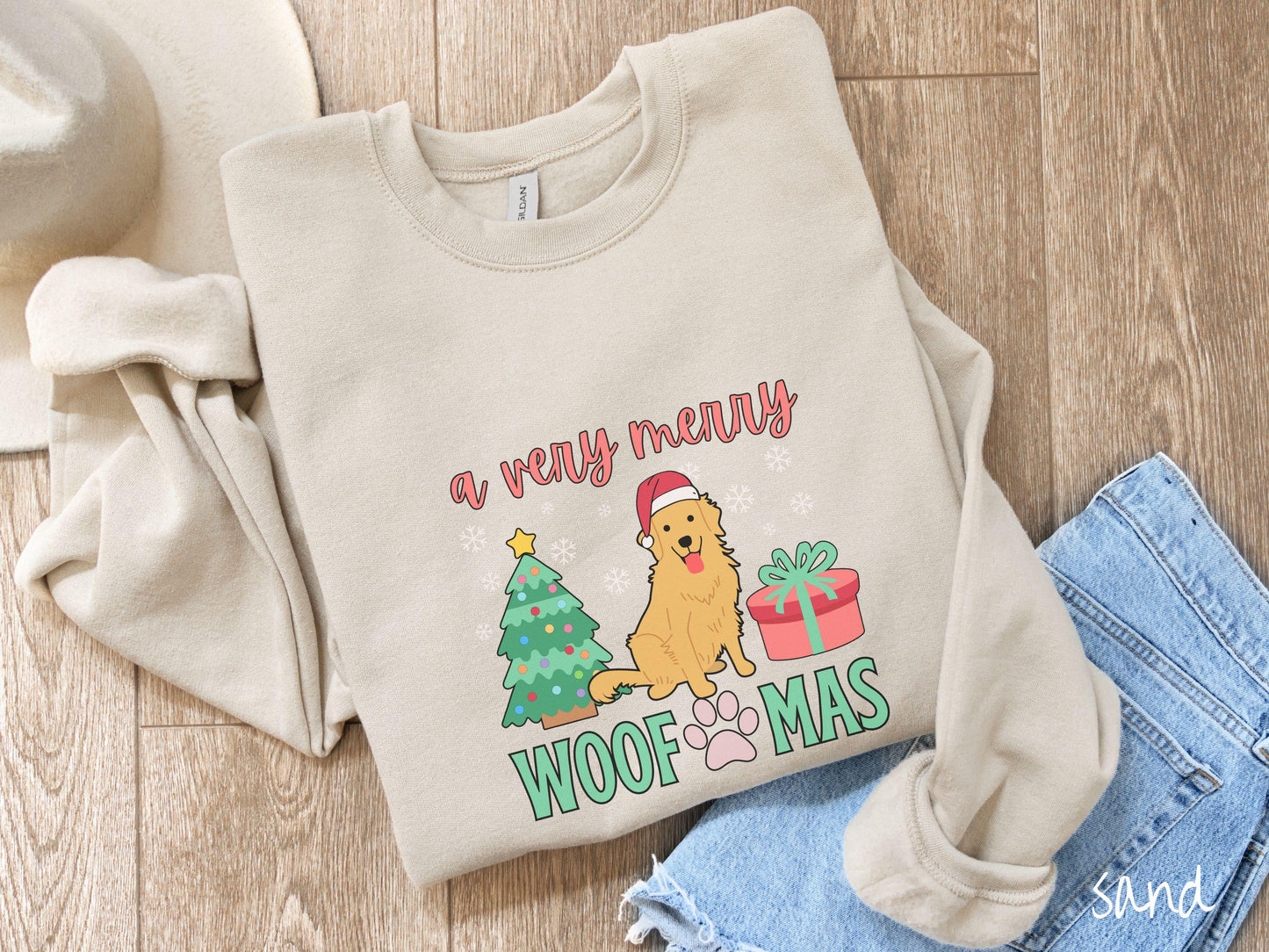 A vintage sand colored Gildan sweatshirt with the text 'a very merry Woofmas' in red and green font, featuring a Golden Retriever wearing a red Santa hat sitting between a Christmas tree and a present with white snowflakes hanging in the air.