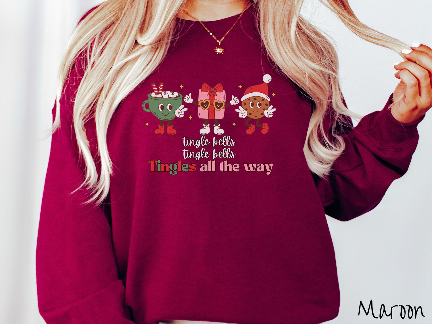 A cute, vintage maroon colored Gildan sweatshirt with text tingle bells, tingle bells, Tingles all the way. Above are a green cup of hot cocoa, a red and pink present wearing heart-shaped glasses, and a chocolate chip cookie wearing a Santa hat.